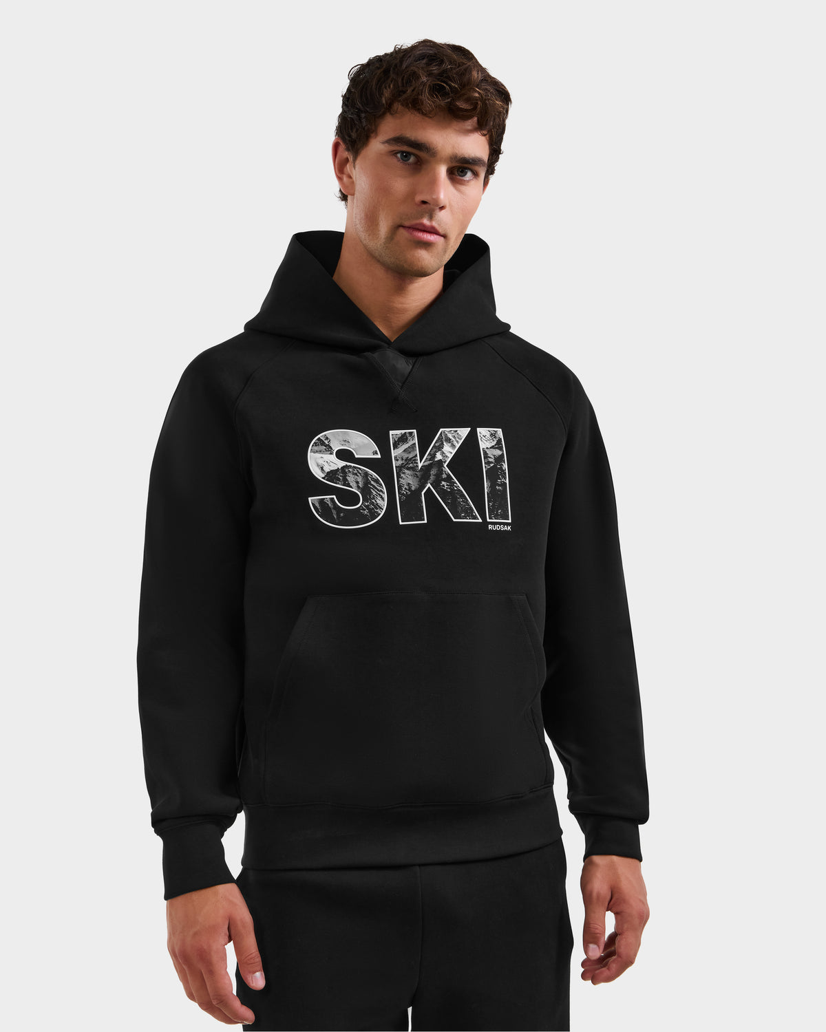 A man wearing the Rudsak MIKAEL - 6525647 Black men’s ski hoodie with a bold SKI logo on the front stands against a light background, looking at the camera with short, curly hair and a neutral expression.