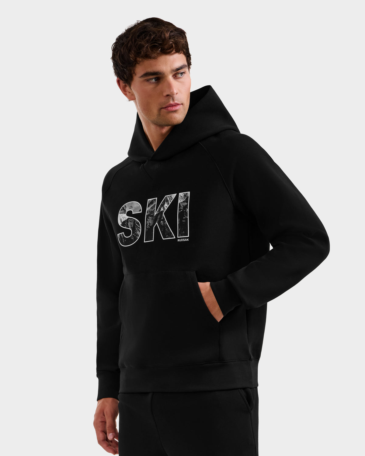 A young man wears the Rudsak MIKAEL - 6525647 Black premium cotton hoodie featuring bold, distressed white SKI lettering on the front—a classic staple of ski culture—standing with one hand in his pocket against a plain background.