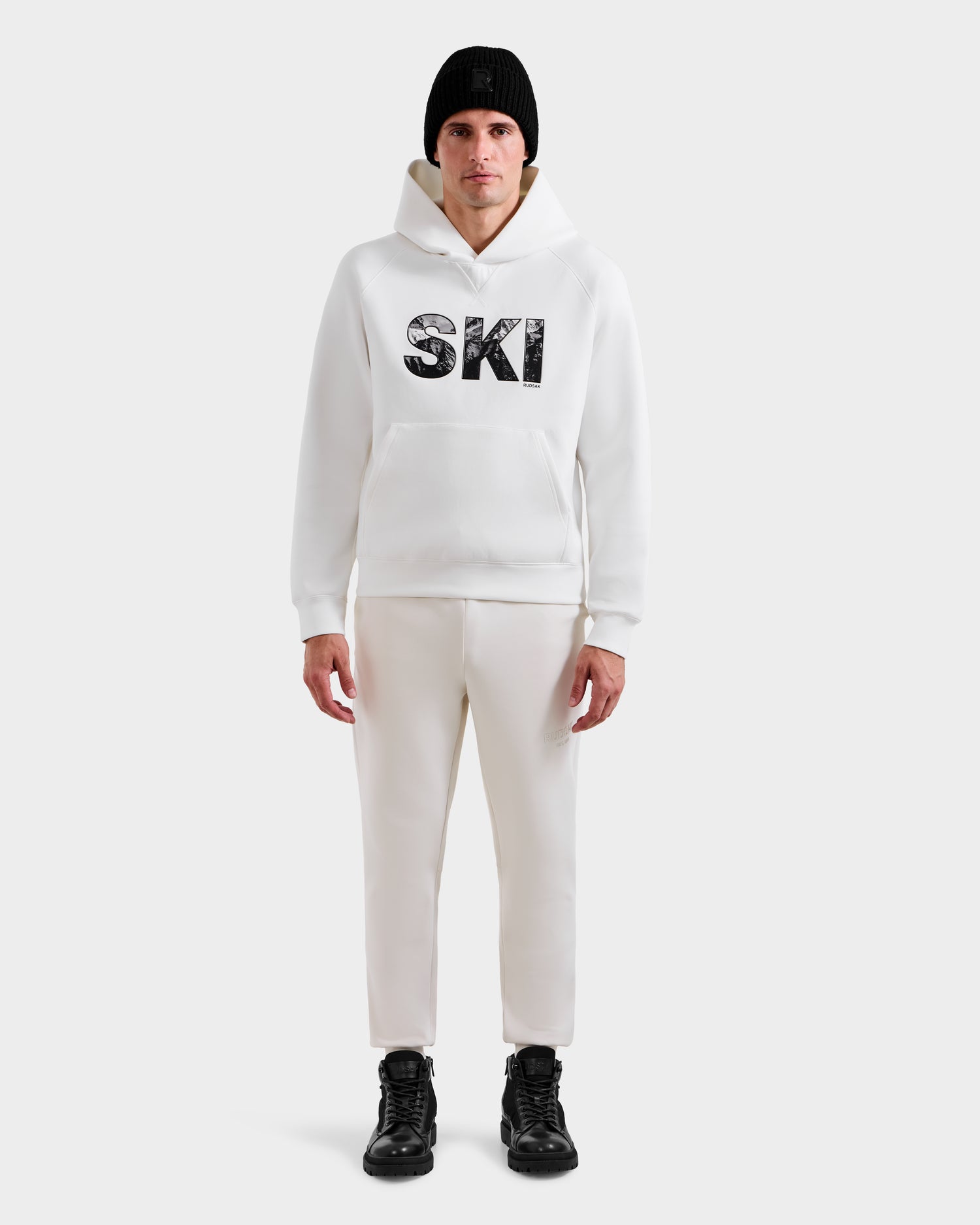 A man faces forward in the Rudsak MIKAEL - 6525647 Iceland men’s ski hoodie with bold SKI lettering, styled with white pants, black boots, and a black beanie against a light gray background—ideal for winter style.