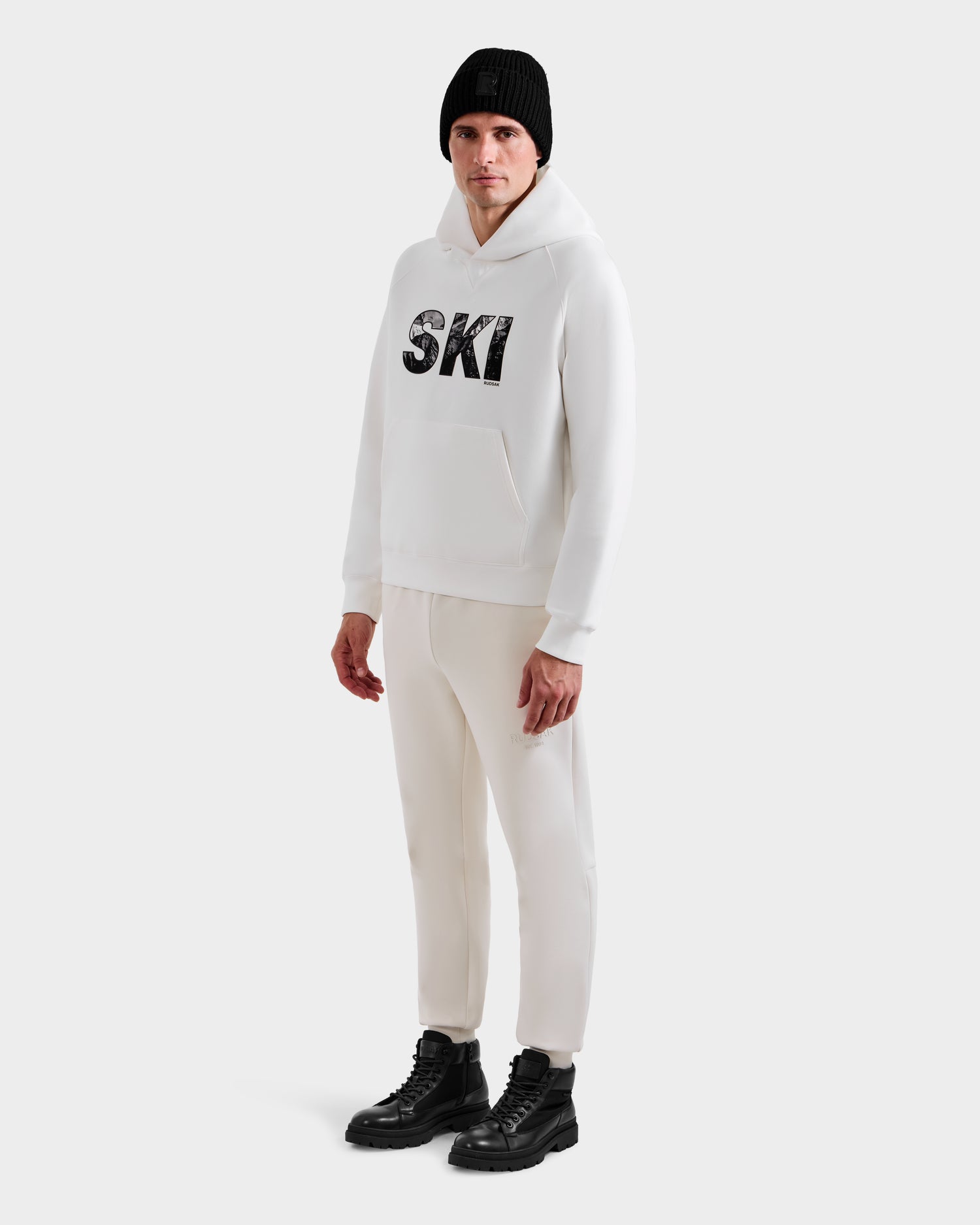 A person wears the Rudsak MIKAEL - 6525647 Iceland men’s ski hoodie with SKI on the front, paired with white pants, black boots, and a black beanie against a plain white background—perfect for any winter wardrobe.