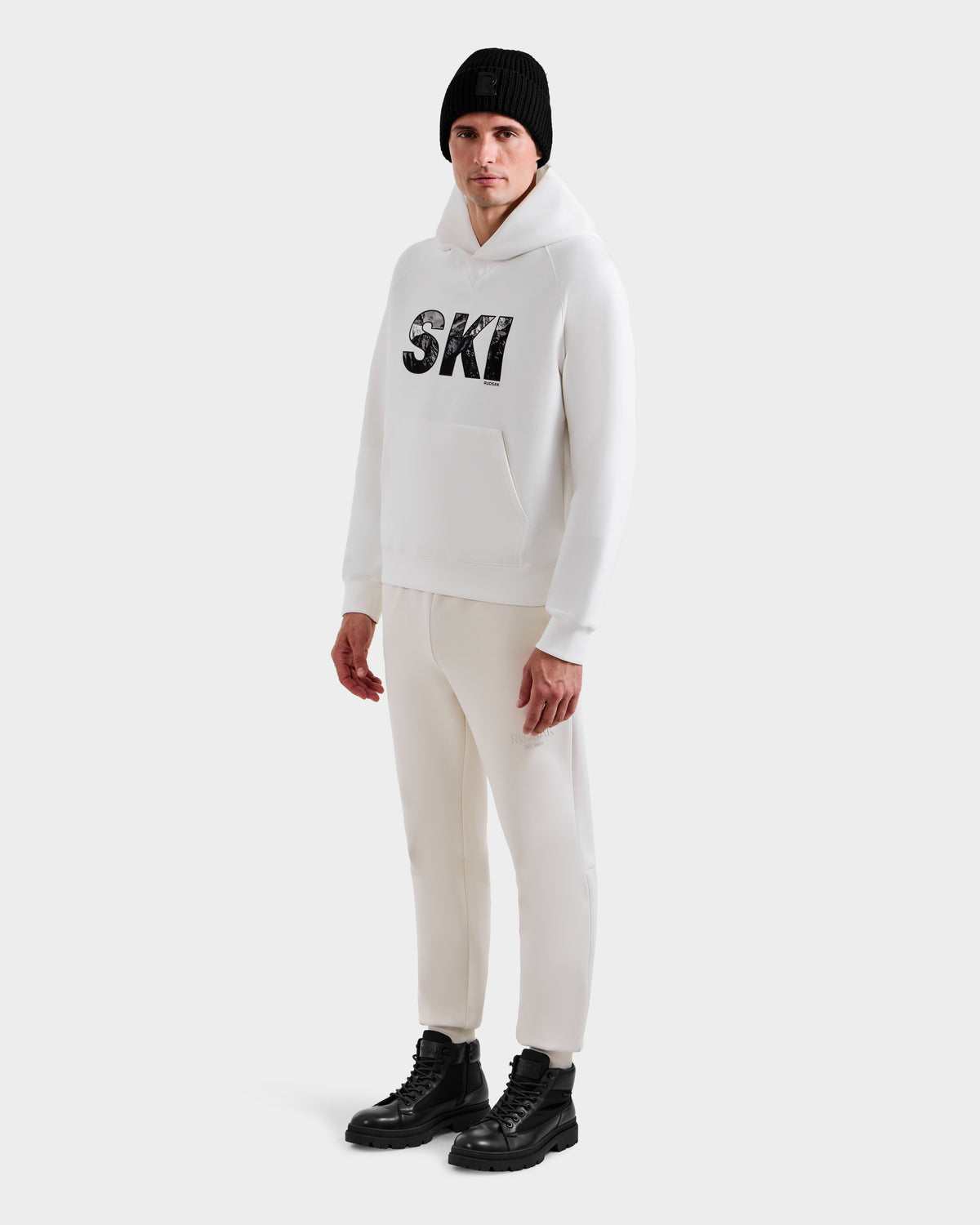 A person wears the Rudsak MIKAEL - 6525647 Iceland men’s ski hoodie with SKI on the front, paired with white pants, black boots, and a black beanie against a plain white background—perfect for any winter wardrobe.