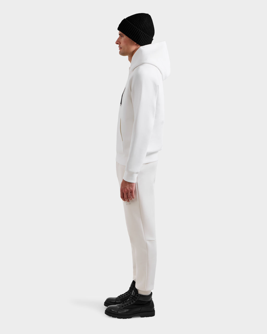 A man stands in profile against a plain background, wearing the Rudsak MIKAEL - 6525647 Iceland ski hoodie with matching white pants, a black beanie, and black boots—an essential winter wardrobe look.