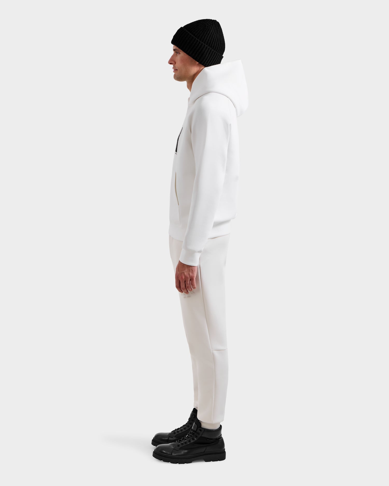 A man stands in profile against a plain background, wearing the Rudsak MIKAEL - 6525647 Iceland ski hoodie with matching white pants, a black beanie, and black boots—an essential winter wardrobe look.