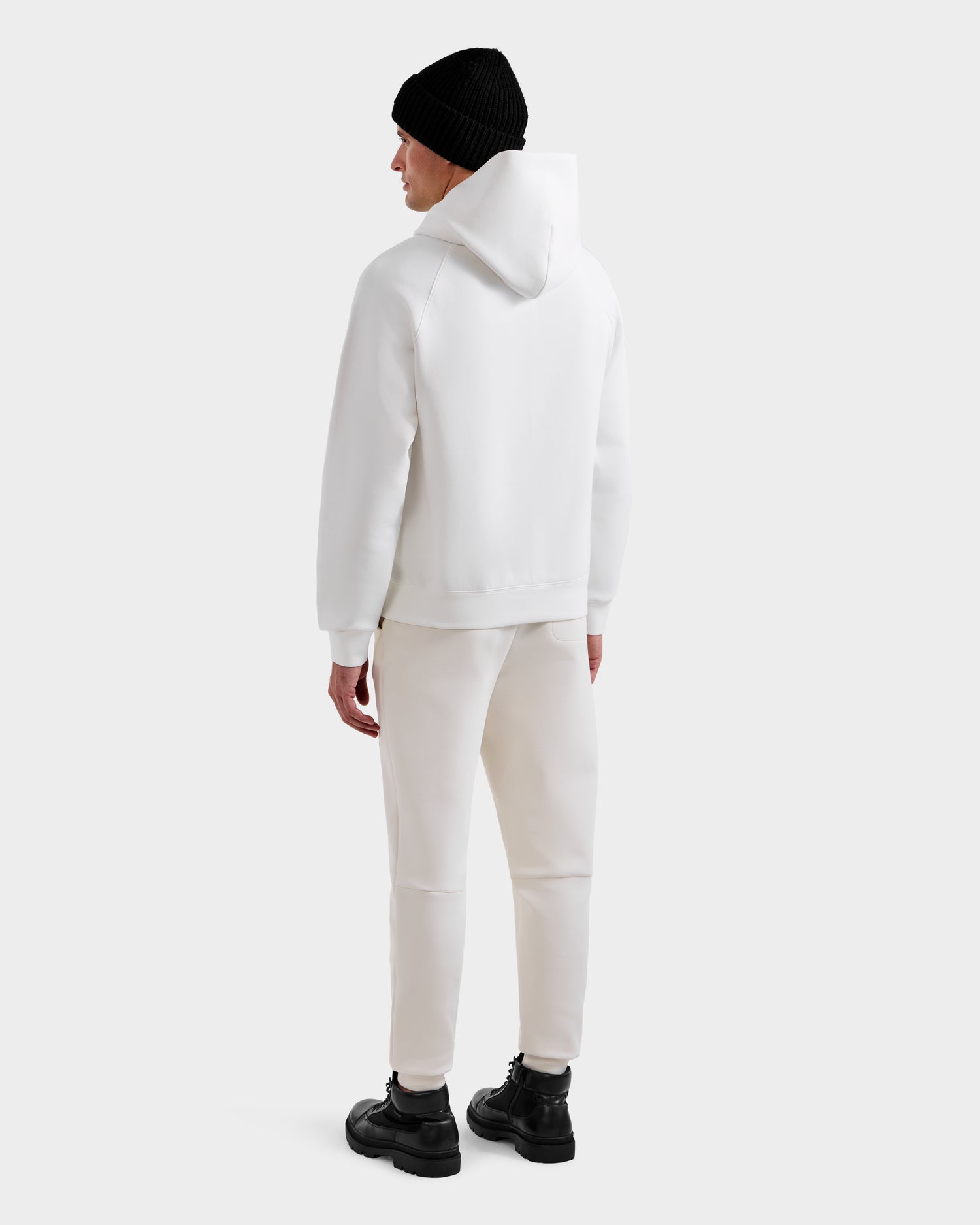A person faces away, wearing the Rudsak MIKAEL - 6525647 Iceland ski hoodie with white pants, a black beanie, and black boots against a plain light background for a stylish winter look.