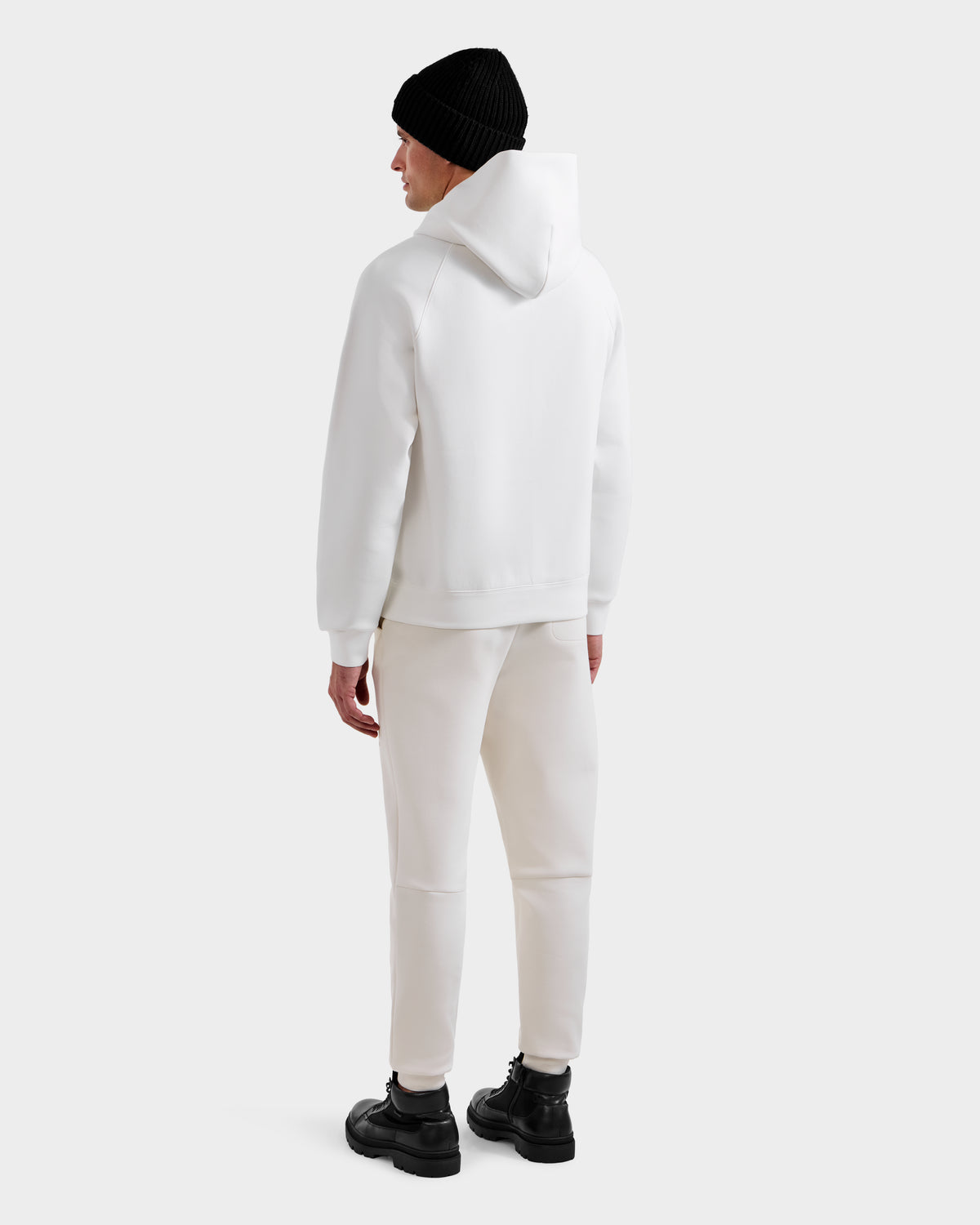 A person faces away, wearing the Rudsak MIKAEL - 6525647 Iceland ski hoodie with white pants, a black beanie, and black boots against a plain light background for a stylish winter look.