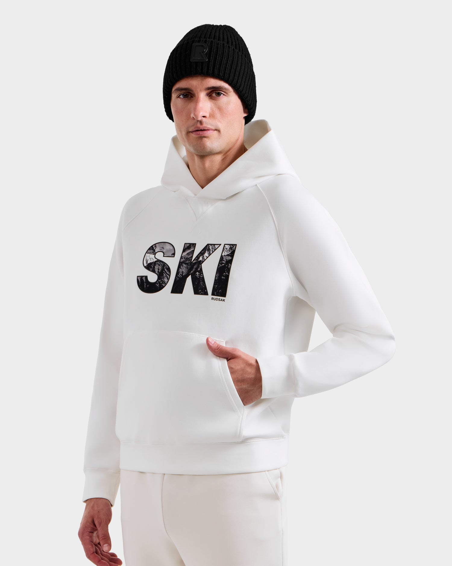 A man wearing the Rudsak MIKAEL - 6525647 Iceland men’s ski hoodie with “SKI” on the front and a black knit beanie, standing with one hand in his pocket against a plain background.