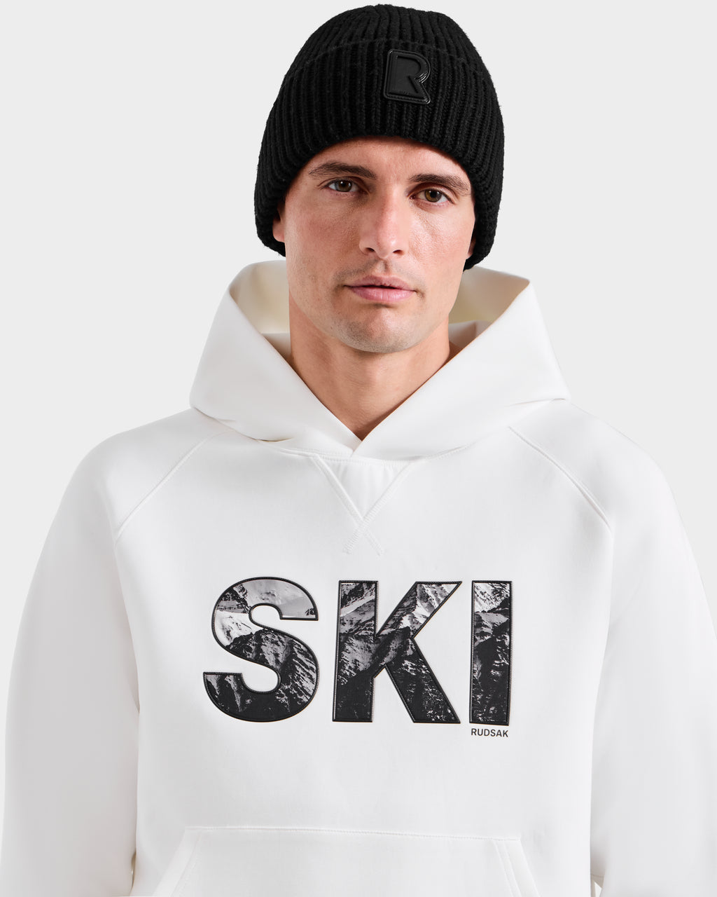 A man wears the Rudsak MIKAEL - 6525647 Iceland ski hoodie with a black knit beanie; "SKI" is boldly printed on the front, making it a must-have addition to any winter wardrobe.