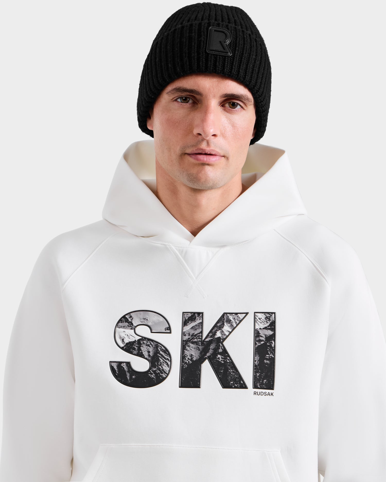 A man wears the Rudsak MIKAEL - 6525647 Iceland ski hoodie with a black knit beanie; "SKI" is boldly printed on the front, making it a must-have addition to any winter wardrobe.