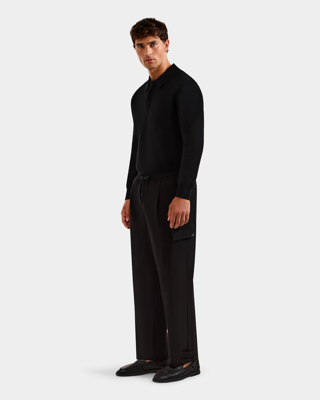 A man stands against a plain background wearing the MIKEY - 6525597 Black long-sleeve shirt by Rudsak, premium men’s trousers with a side pocket, and black shoes, looking slightly to the side with a neutral expression.