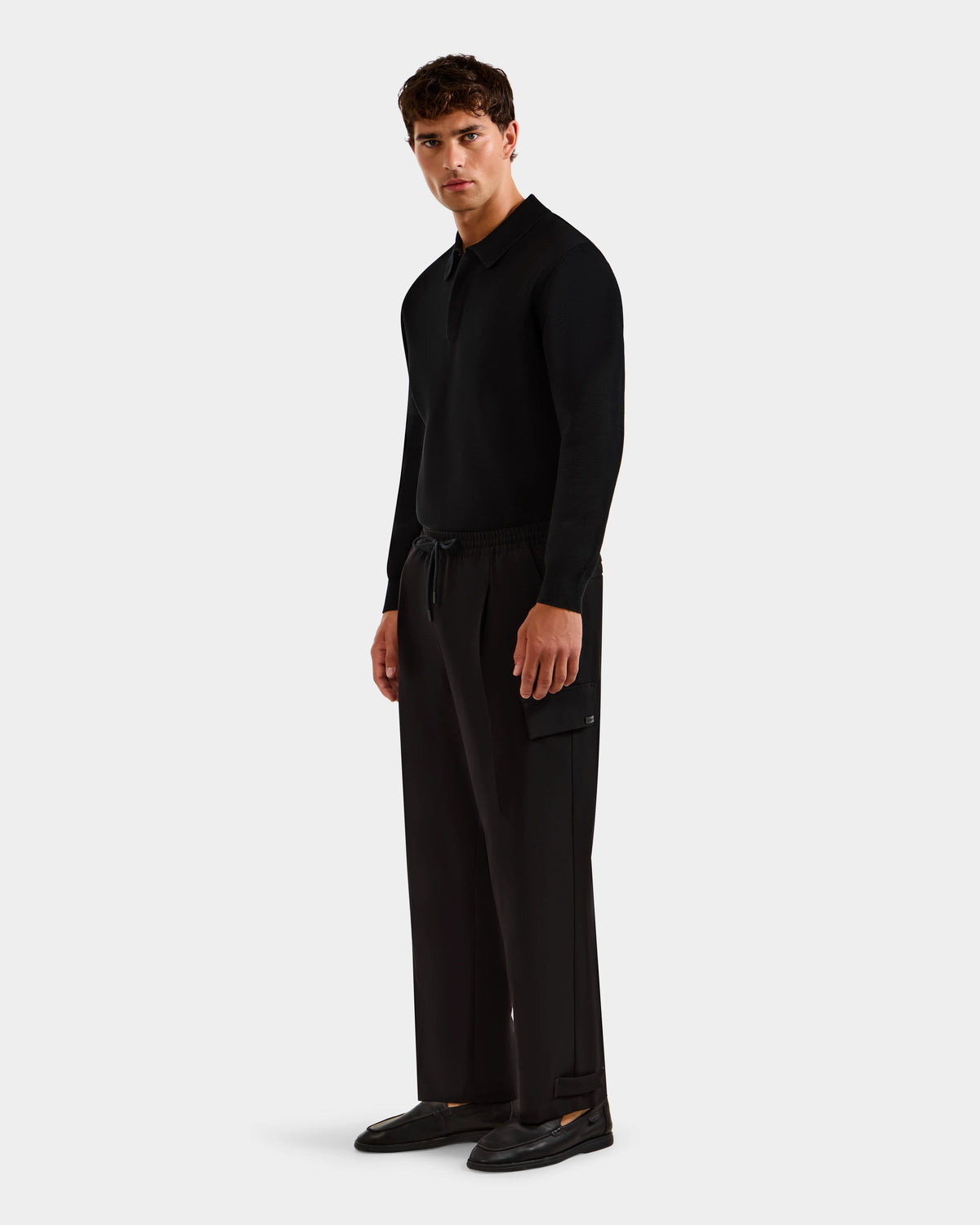 A man stands against a plain background wearing the MIKEY - 6525597 Black long-sleeve shirt by Rudsak, premium men’s trousers with a side pocket, and black shoes, looking slightly to the side with a neutral expression.