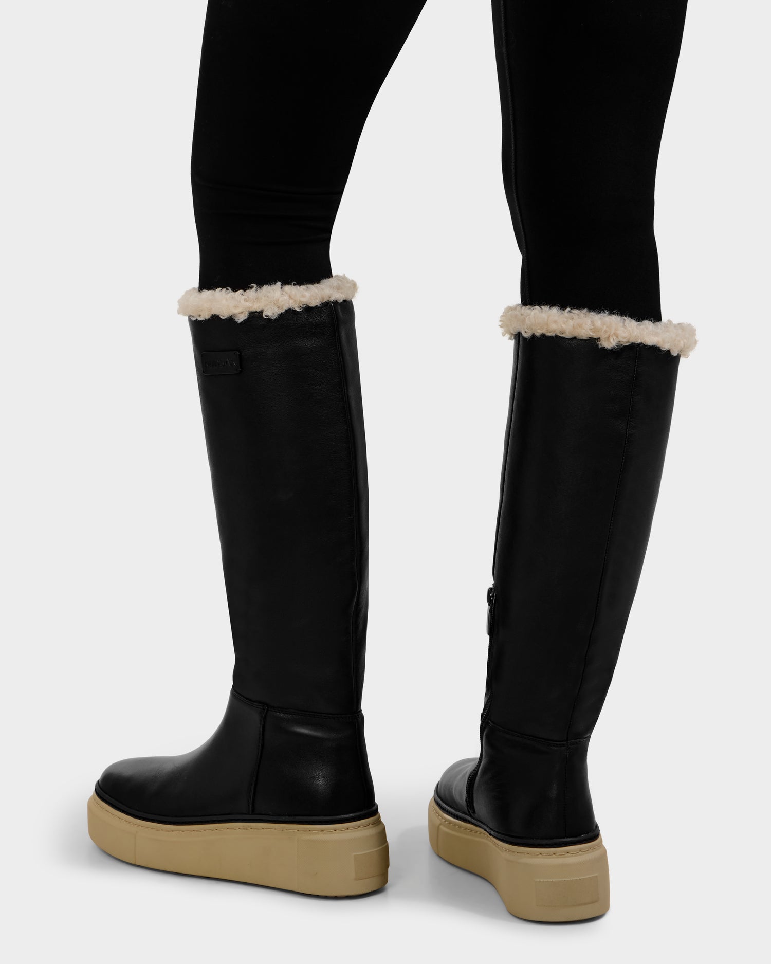 A person wearing Rudsak MIKIE L - 8225580 Black boots with beige soles and white fleece trim, paired with black leggings, stands on a light-colored surface.