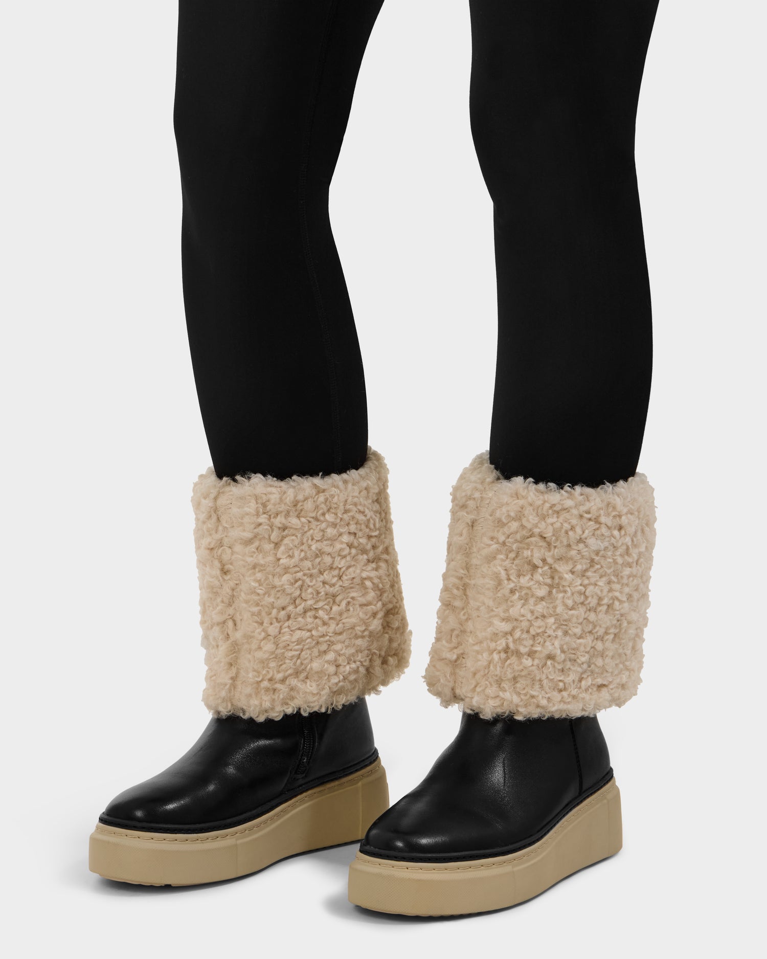 A person wears black leggings and Rudsak MIKIE L - 8225580 Black foldover shearling boots with thick, cream-colored fuzzy cuffs. The background is plain white.