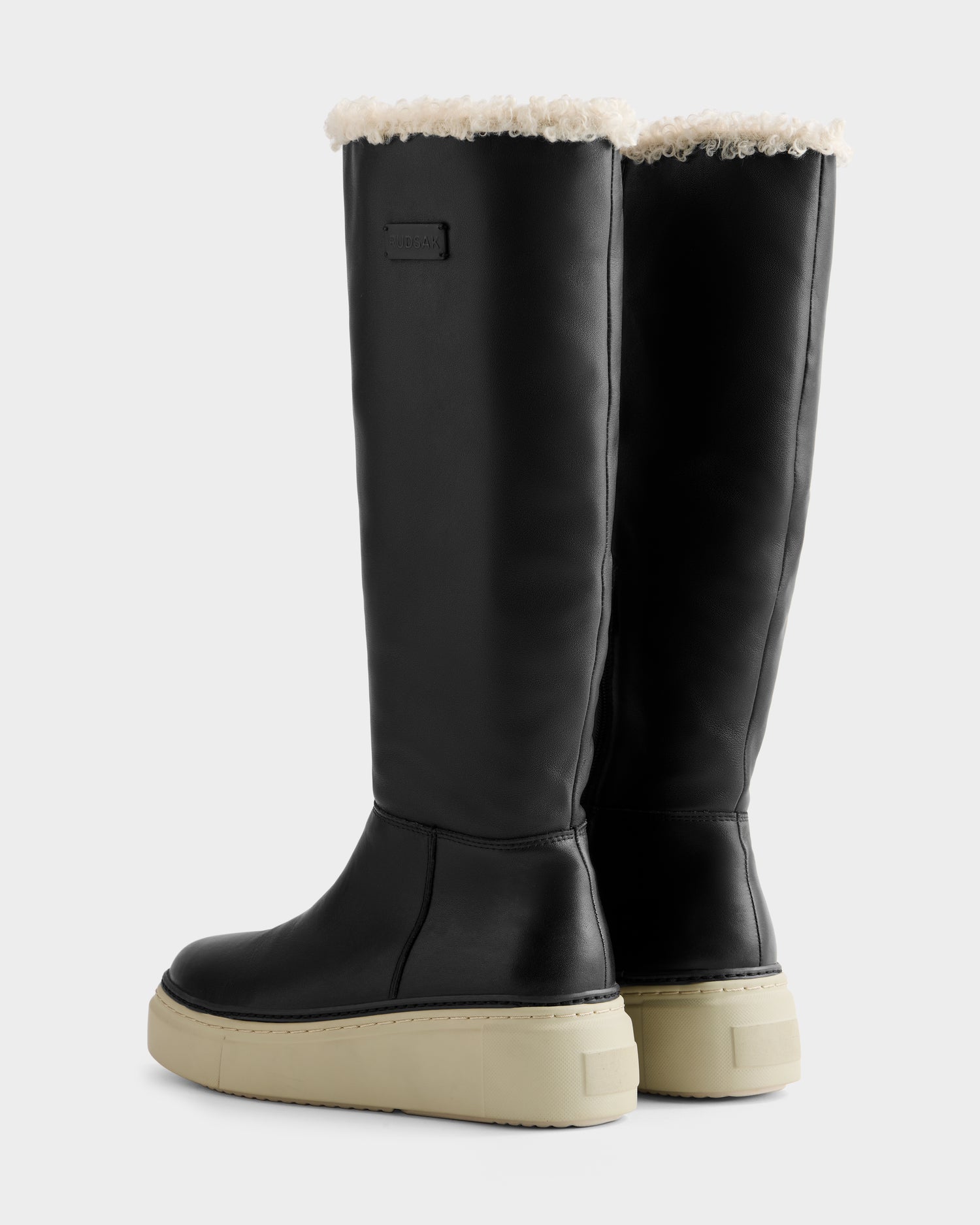 The MIKIE L - 8225580 Black boots by Rudsak feature black knee-high uppers, cream chunky soles, and white shearling trim at the top, shown against a plain light background.