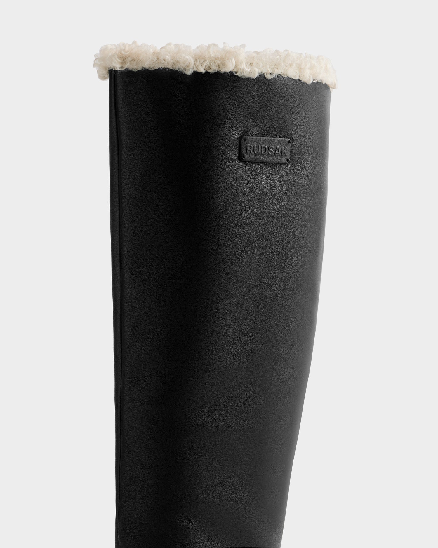 A close-up of the Rudsak MIKIE L - 8225580 Black tall leather boot with a fuzzy white lining at the top and a visible brand logo patch on the upper calf.