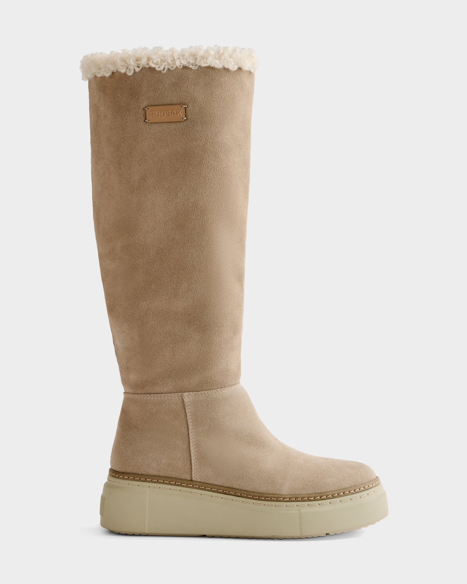 The Rudsak MIKIE - 8225579 Almond is a beige knee-high suede boot featuring a thick light tan sole, white shearling trim at the top, stitched detailing around the sole, and a small rectangular logo patch near the top.