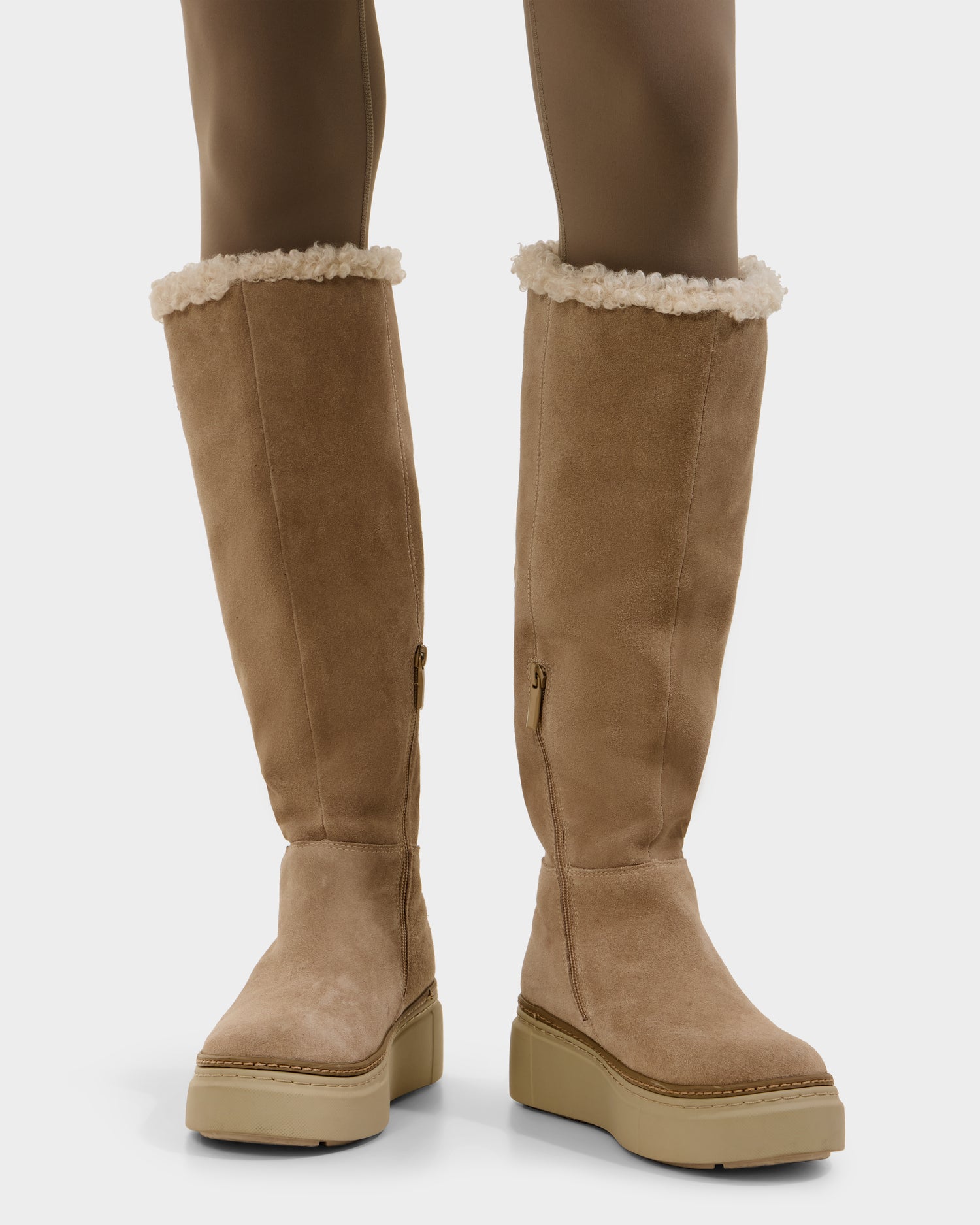 A person wears Rudsak’s MIKIE - 8225579 Almond tan suede knee-high boots with light shearling trim, side zipper, and thick rubber soles, paired with brown tights—ideal cold-weather foldover boots.