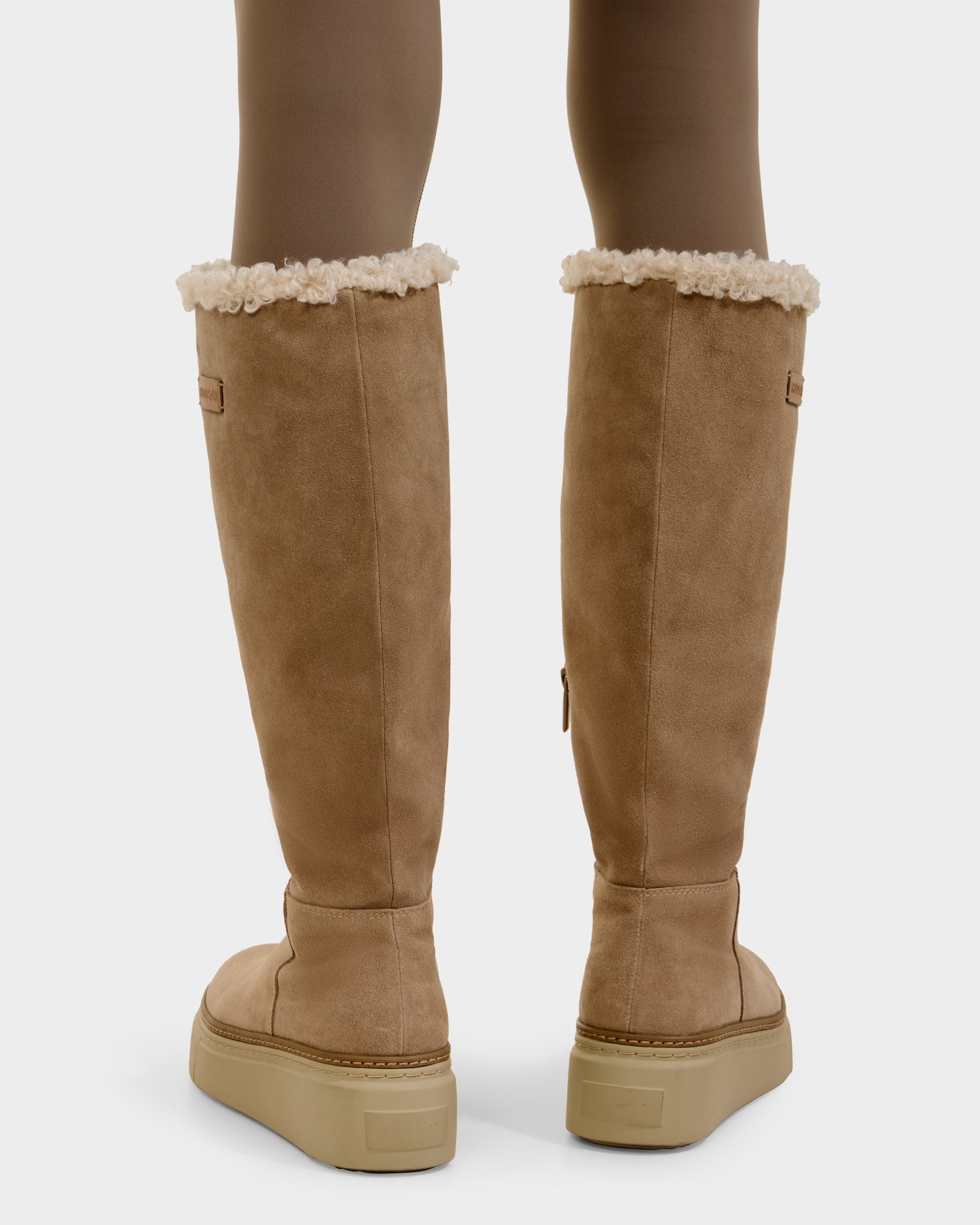 A person stands on a white background wearing Rudsak MIKIE - 8225579 Almond women's suede boots with cream shearling trim, flat light tan rubber soles, and foldover shearling at the top. Only their lower legs are visible.