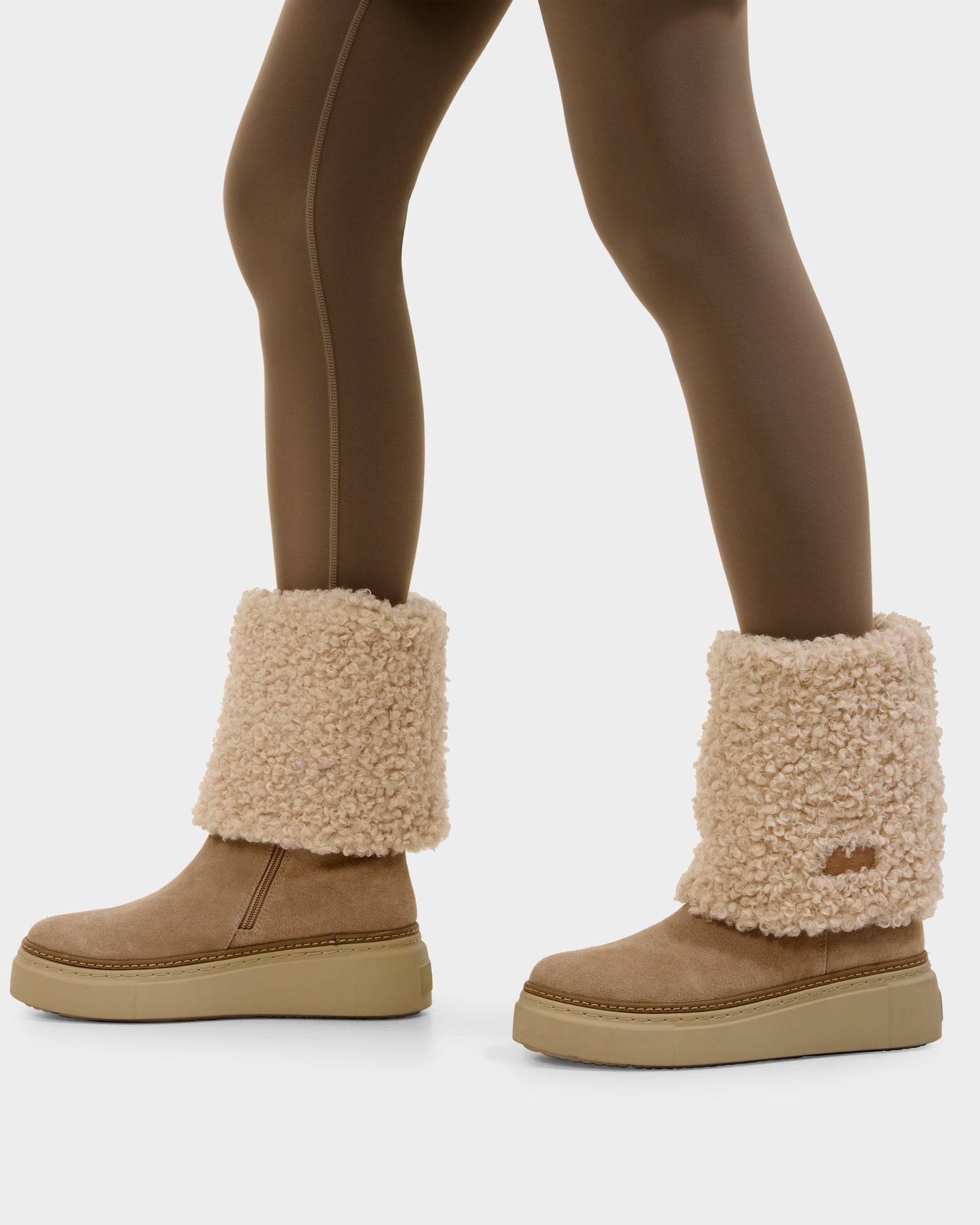 A person wears brown leggings and Rudsak MIKIE - 8225579 Almond women’s suede boots with thick shearling foldover cuffs and a flat platform sole, highlighting the cozy, textured style. The image centers on their lower legs and feet.