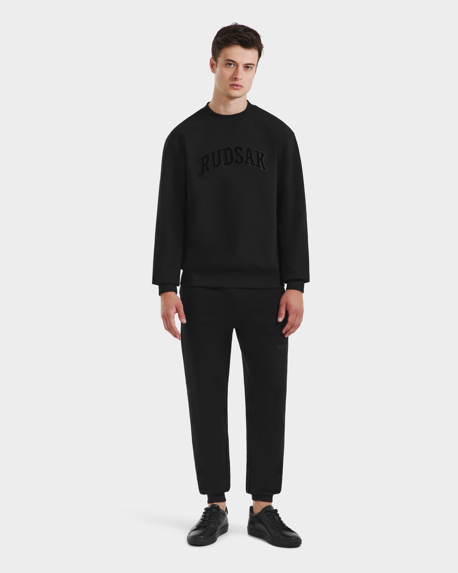 A young man stands against a plain background, wearing the Rudsak MILOS - 6525646 Black men’s premium crewneck sweatshirt and pants set with RUDSAK across the chest, paired with black sneakers.