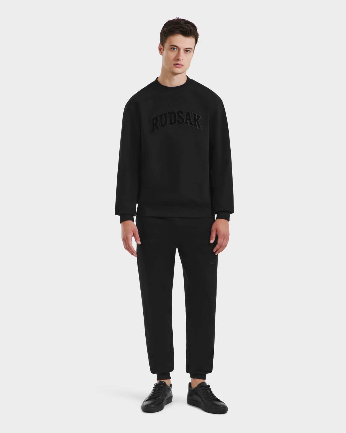 A young man stands against a plain background, wearing the Rudsak MILOS - 6525646 Black men’s premium crewneck sweatshirt and pants set with RUDSAK across the chest, paired with black sneakers.