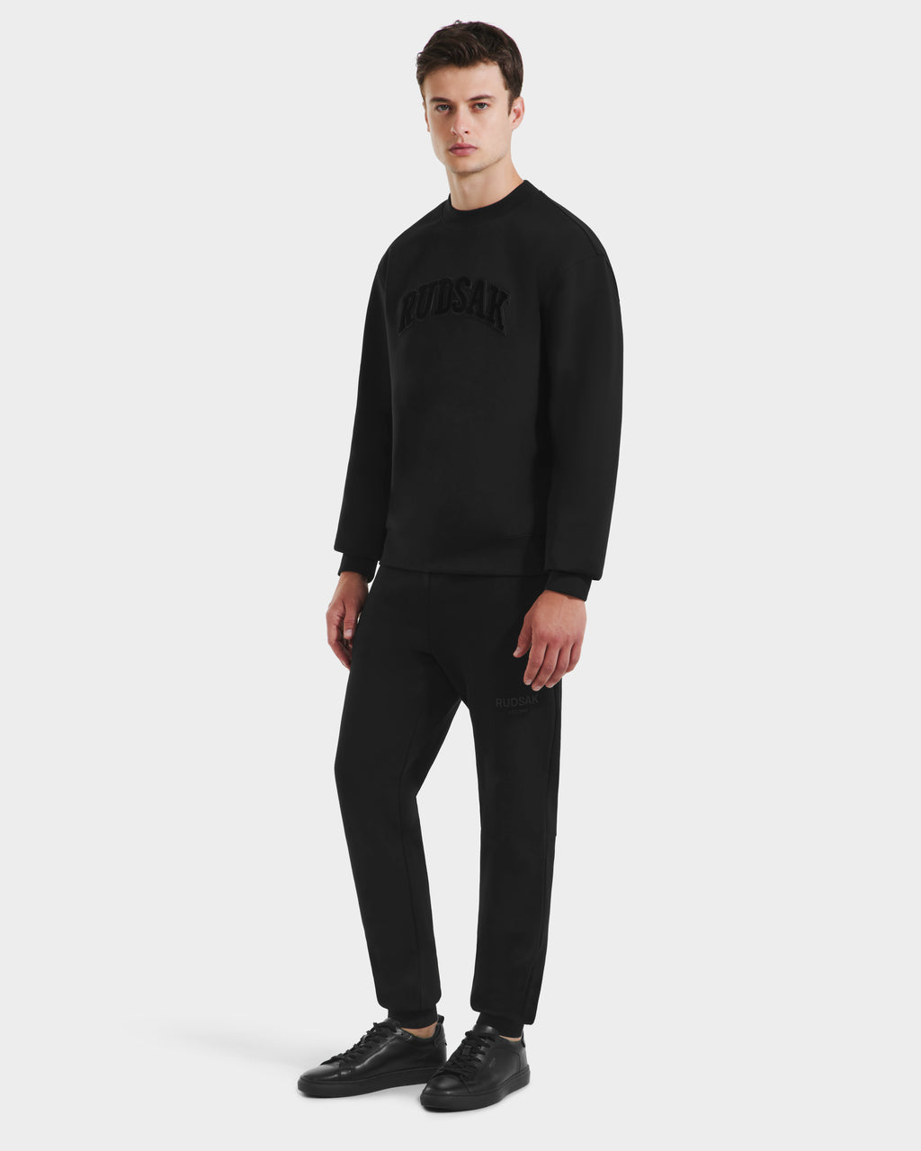 A man stands against a white background in minimalist style, wearing Rudsaks MILOS - 6525646 Black mens premium cotton-blend crewneck sweatshirt with matching joggers and black sneakers for a sleek, comfortable look.