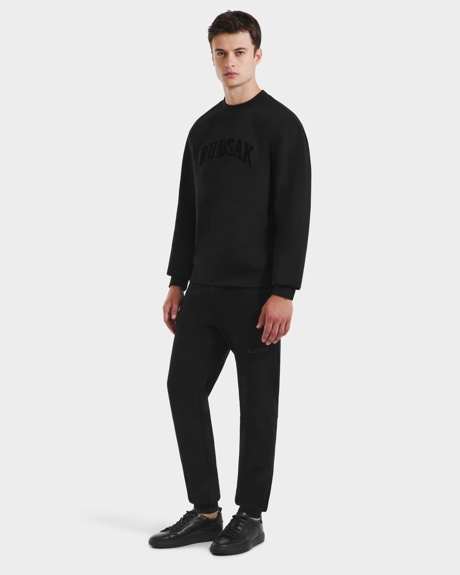 A man stands against a white background in minimalist style, wearing Rudsaks MILOS - 6525646 Black mens premium cotton-blend crewneck sweatshirt with matching joggers and black sneakers for a sleek, comfortable look.