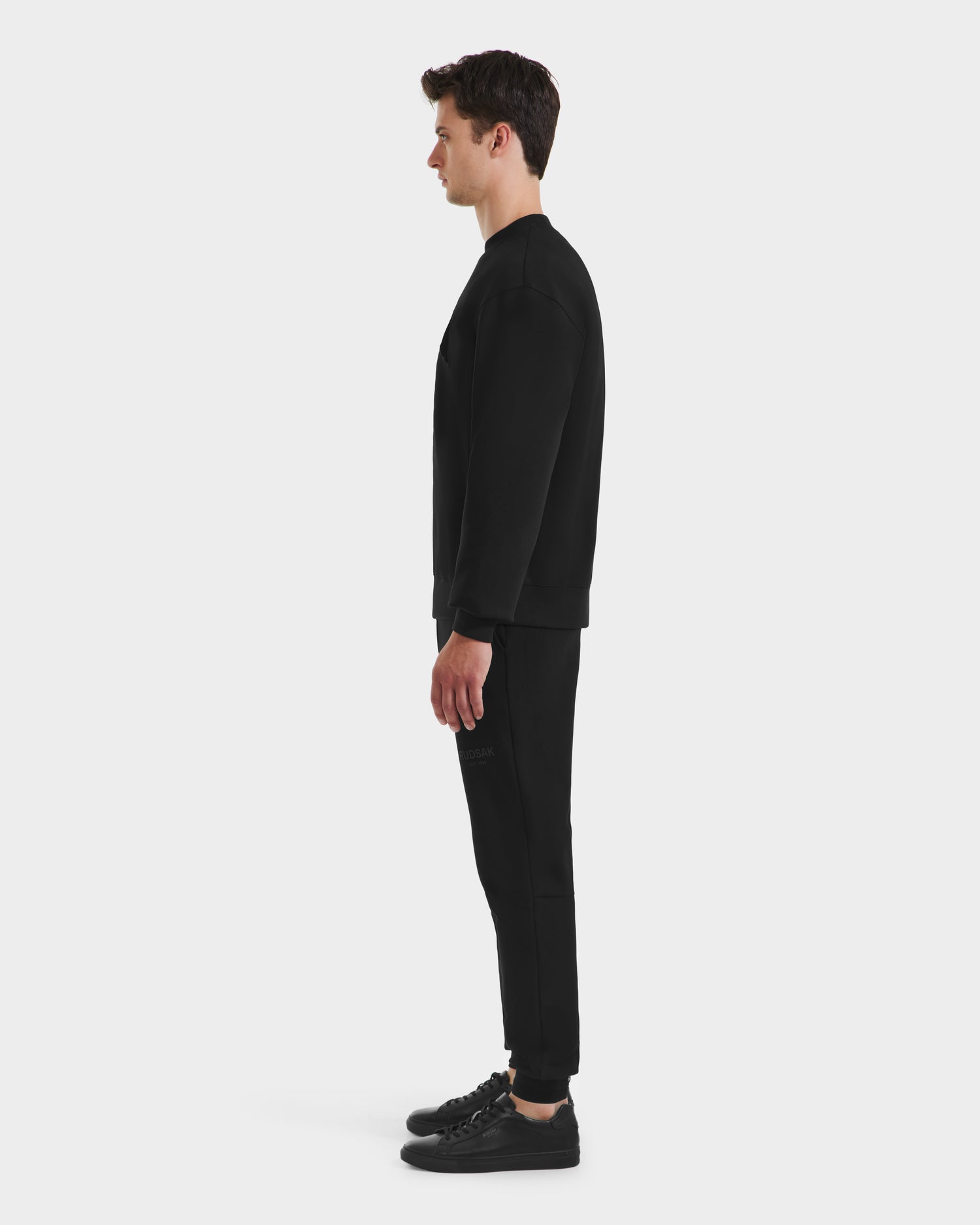 A man in profile, facing left, wears the minimalist all-black MILOS - 6525646 Black sweatshirt and joggers by Rudsak with sneakers, set against a plain white background.