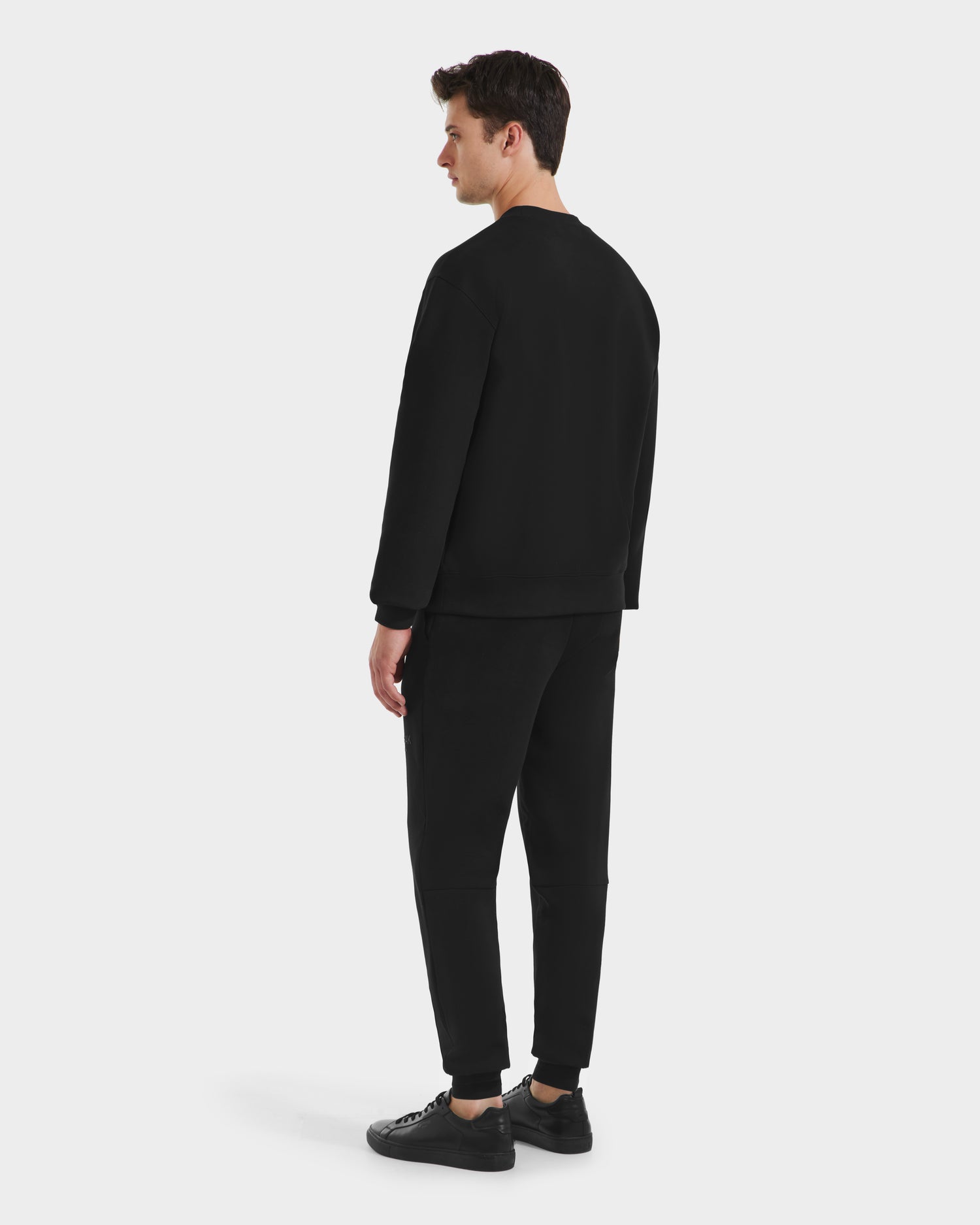 A man stands facing away, wearing the Rudsak MILOS - 6525646 Black men’s premium crewneck sweatshirt, black pants, and black sneakers. The minimalist look is set against a plain white background.