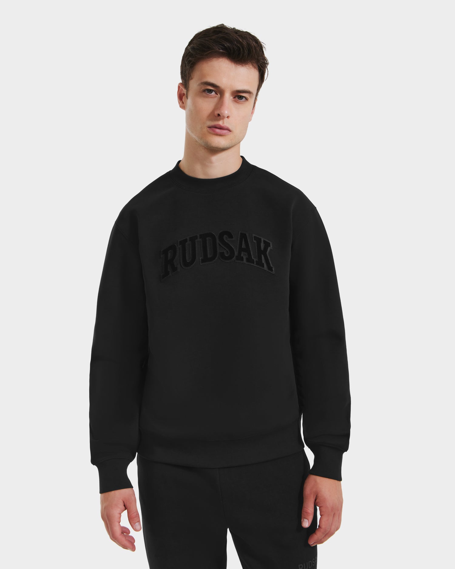 A young man stands against a plain white background, wearing the Rudsak MILOS - 6525646 Black men’s premium cotton-blend crewneck sweatshirt with bold black chest lettering, paired with matching black pants.