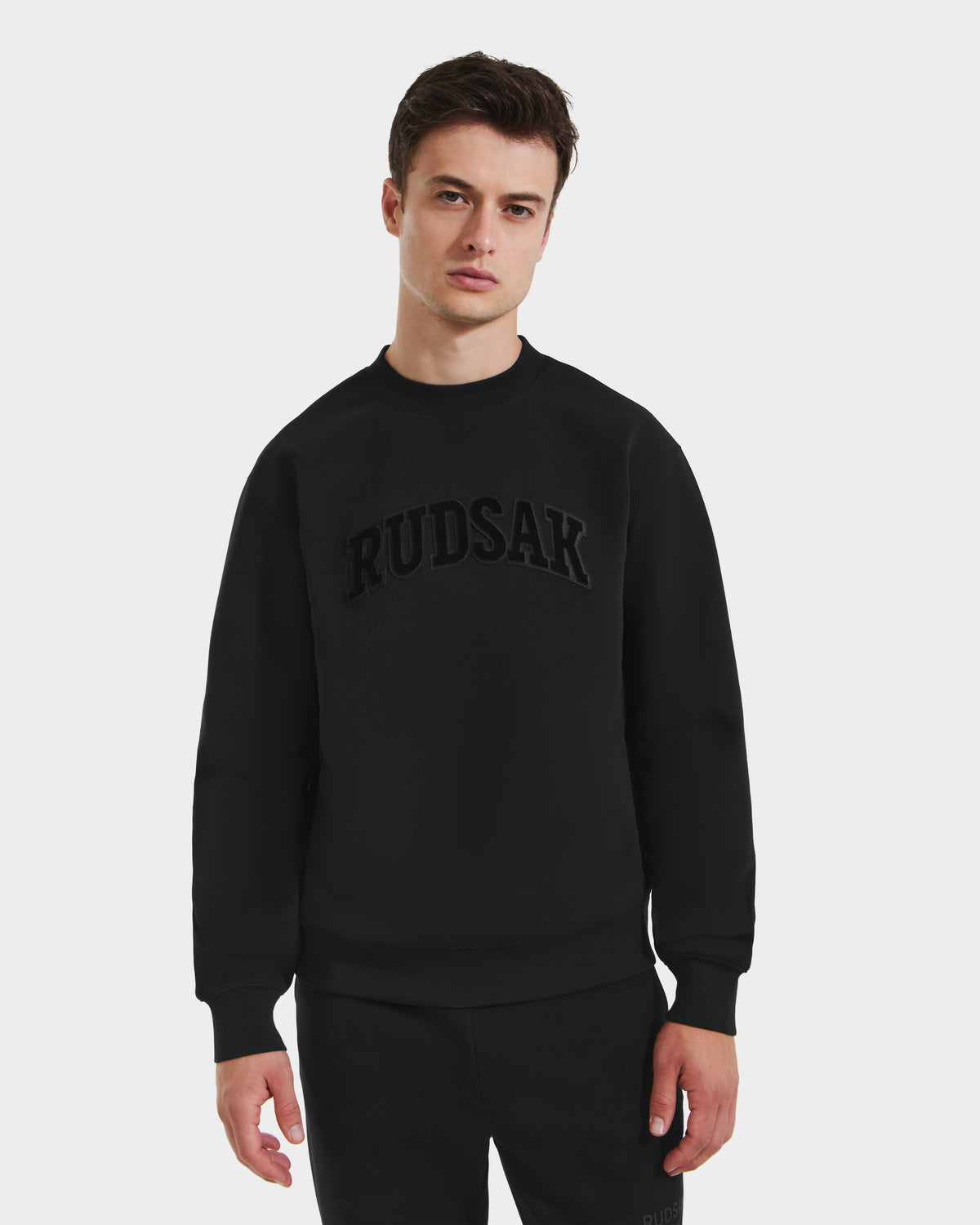 A young man stands against a plain white background, wearing the Rudsak MILOS - 6525646 Black men’s premium cotton-blend crewneck sweatshirt with bold black chest lettering, paired with matching black pants.