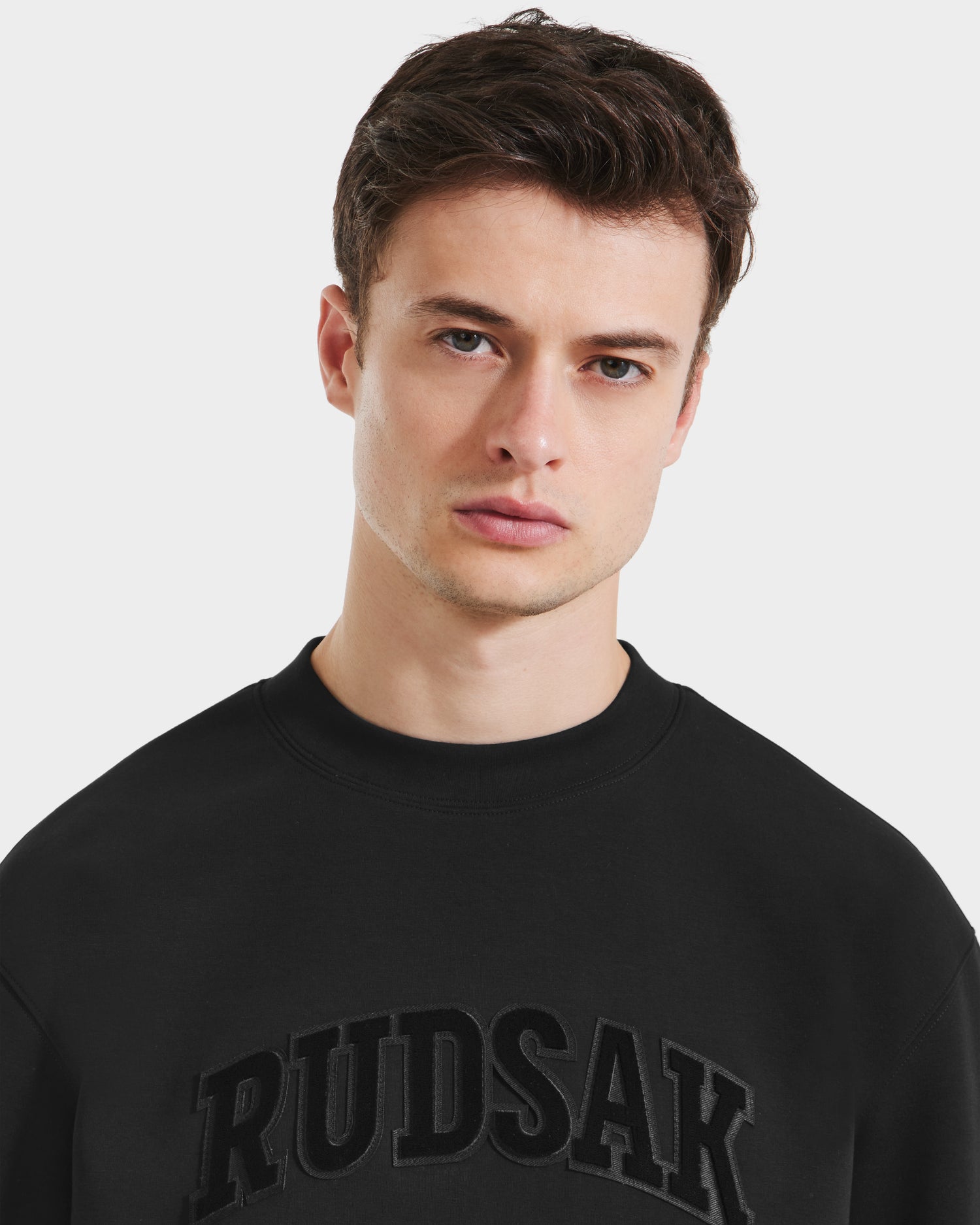 A young man with short brown hair models the Rudsak MILOS - 6525646 Black men’s premium crewneck sweatshirt, featuring bold RUDSAK lettering across the chest, against a plain white background for a minimalist style.