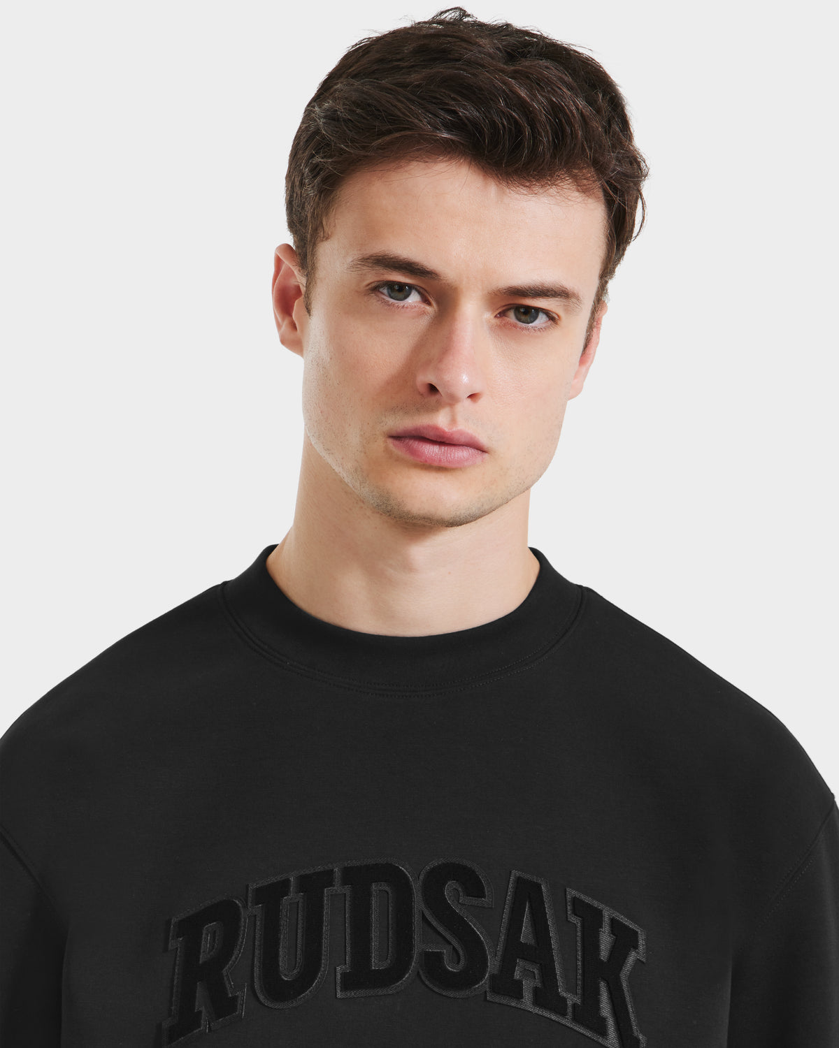 A young man with short brown hair models the Rudsak MILOS - 6525646 Black men’s premium crewneck sweatshirt, featuring bold RUDSAK lettering across the chest, against a plain white background for a minimalist style.