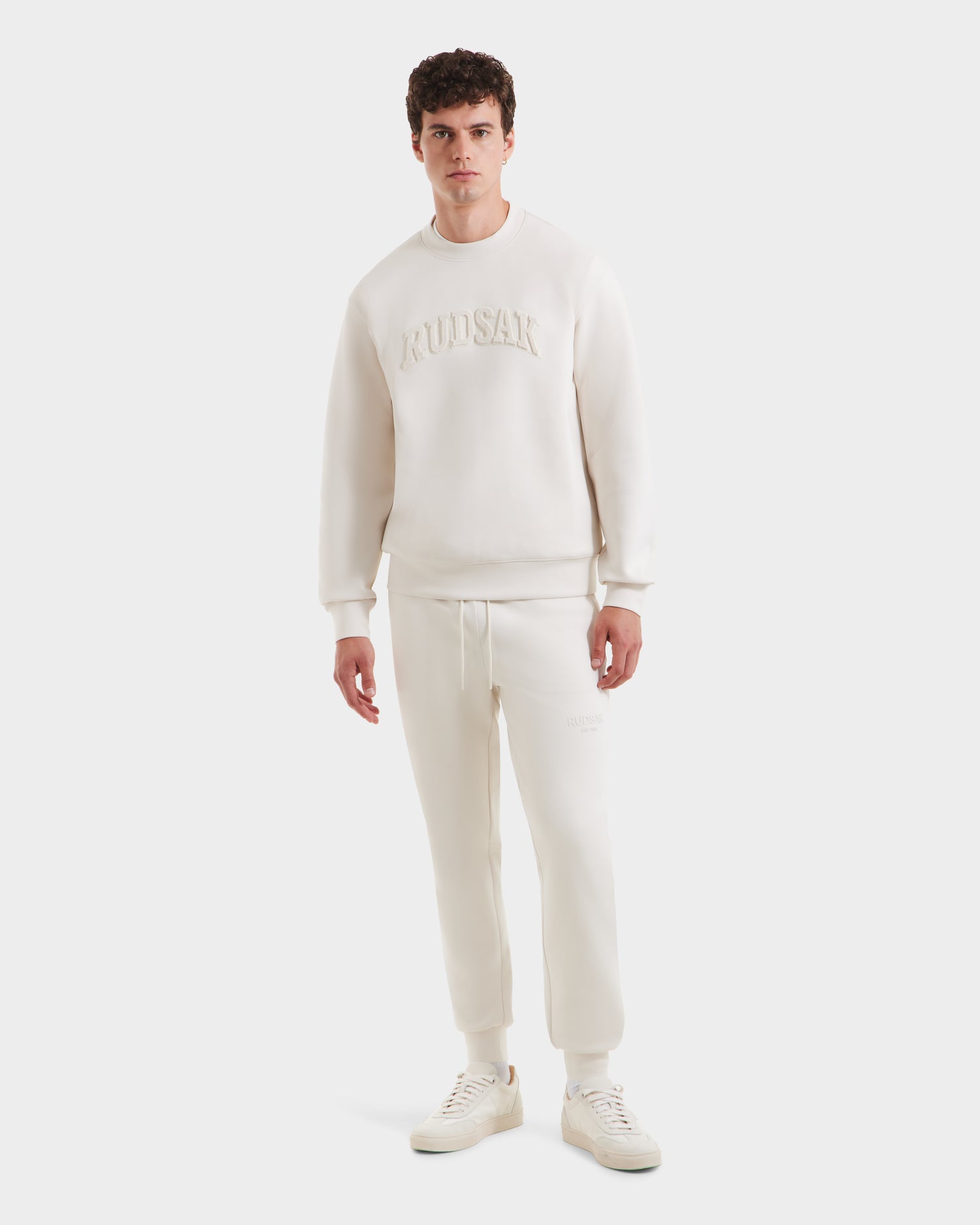 A man faces forward in a minimalist style, wearing Rudsak’s MILOS - 6525646 Vanilla men’s crewneck sweatshirt and jogger pants in off-white, featuring embossed branding, paired with white sneakers against a plain white background.