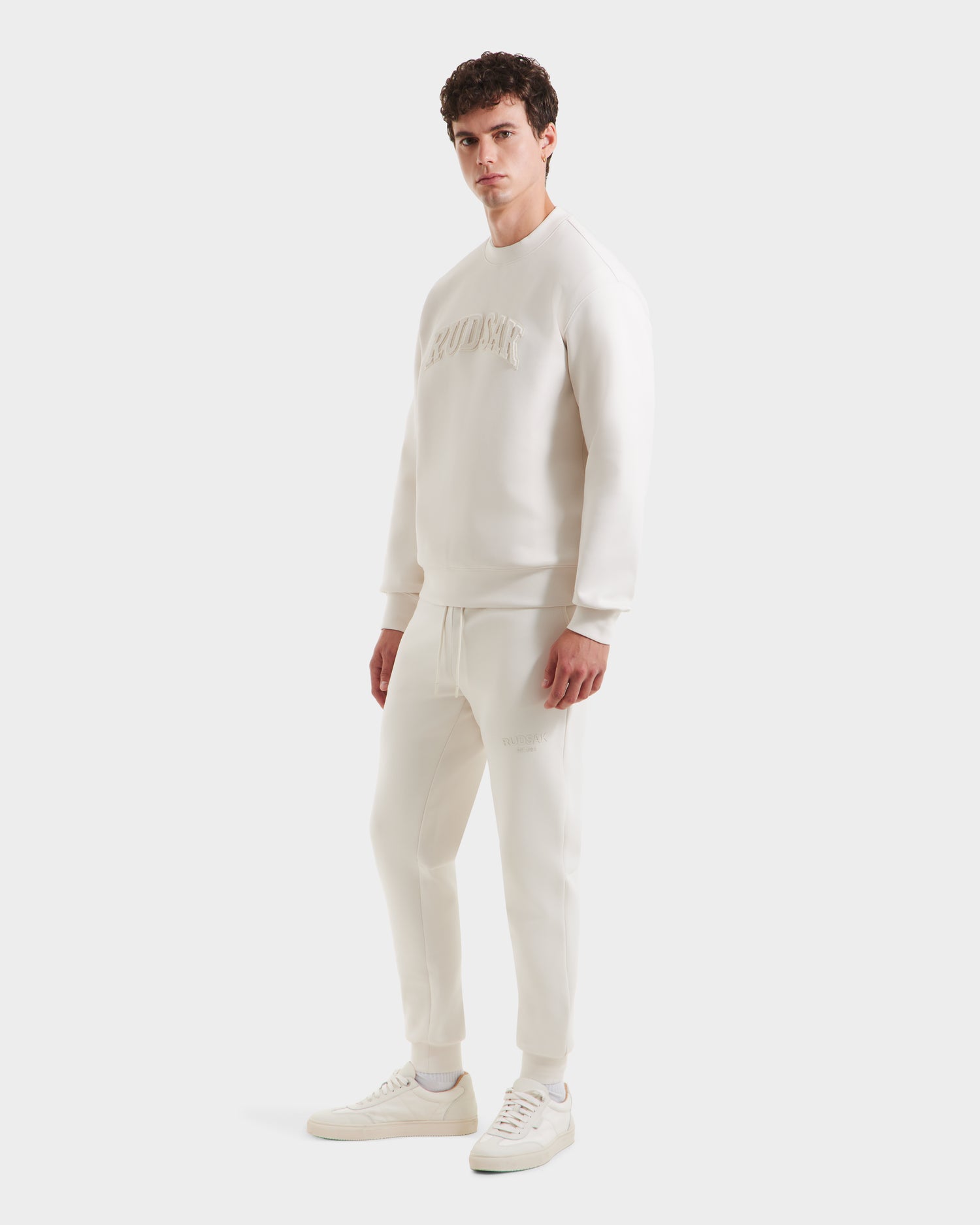 A man stands facing forward in a minimalist style, wearing the Rudsak MILOS - 6525646 Vanilla men’s crewneck sweatshirt with matching joggers and white sneakers against a plain white background.