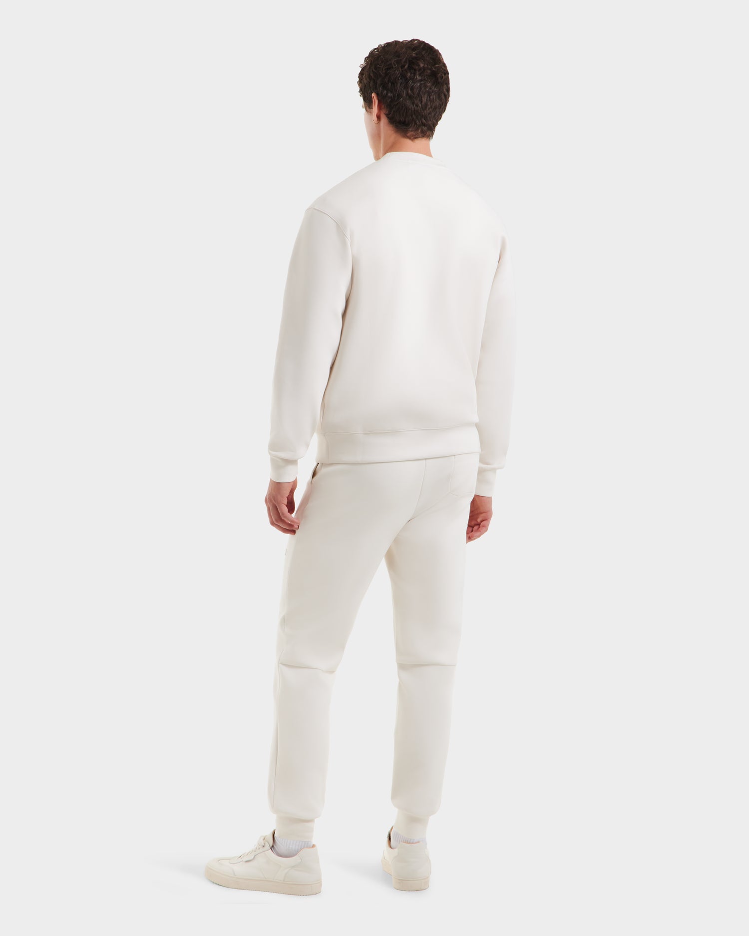 A man, seen from behind, wears Rudsak’s MILOS - 6525646 Vanilla crewneck sweatshirt and sweatpants in a premium cotton blend with white sneakers, styled minimally against a plain white background.