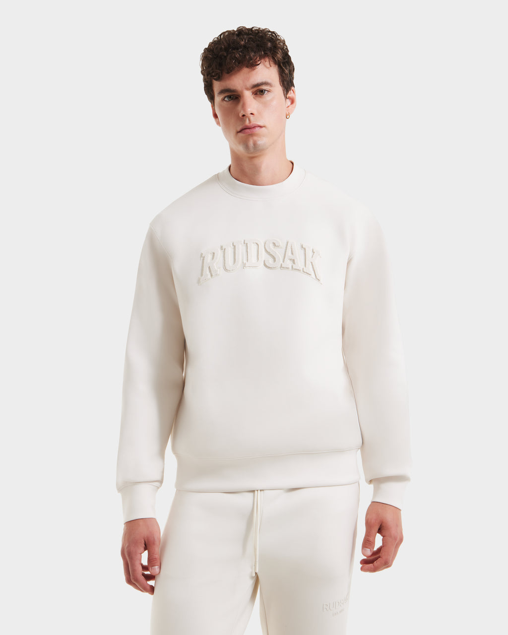 A young man with curly hair wears the Rudsak MILOS - 6525646 Vanilla men’s crewneck sweatshirt, crafted from a premium cotton blend and embossed with Rudsak on the chest, standing against a white background and looking at the camera.