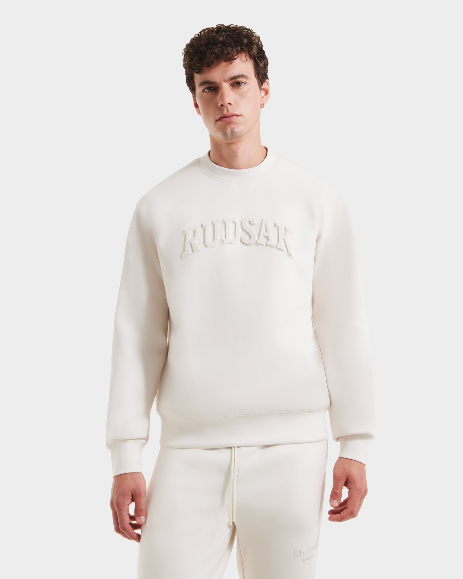 A young man with curly hair wears the Rudsak MILOS - 6525646 Vanilla men’s crewneck sweatshirt, crafted from a premium cotton blend and embossed with Rudsak on the chest, standing against a white background and looking at the camera.