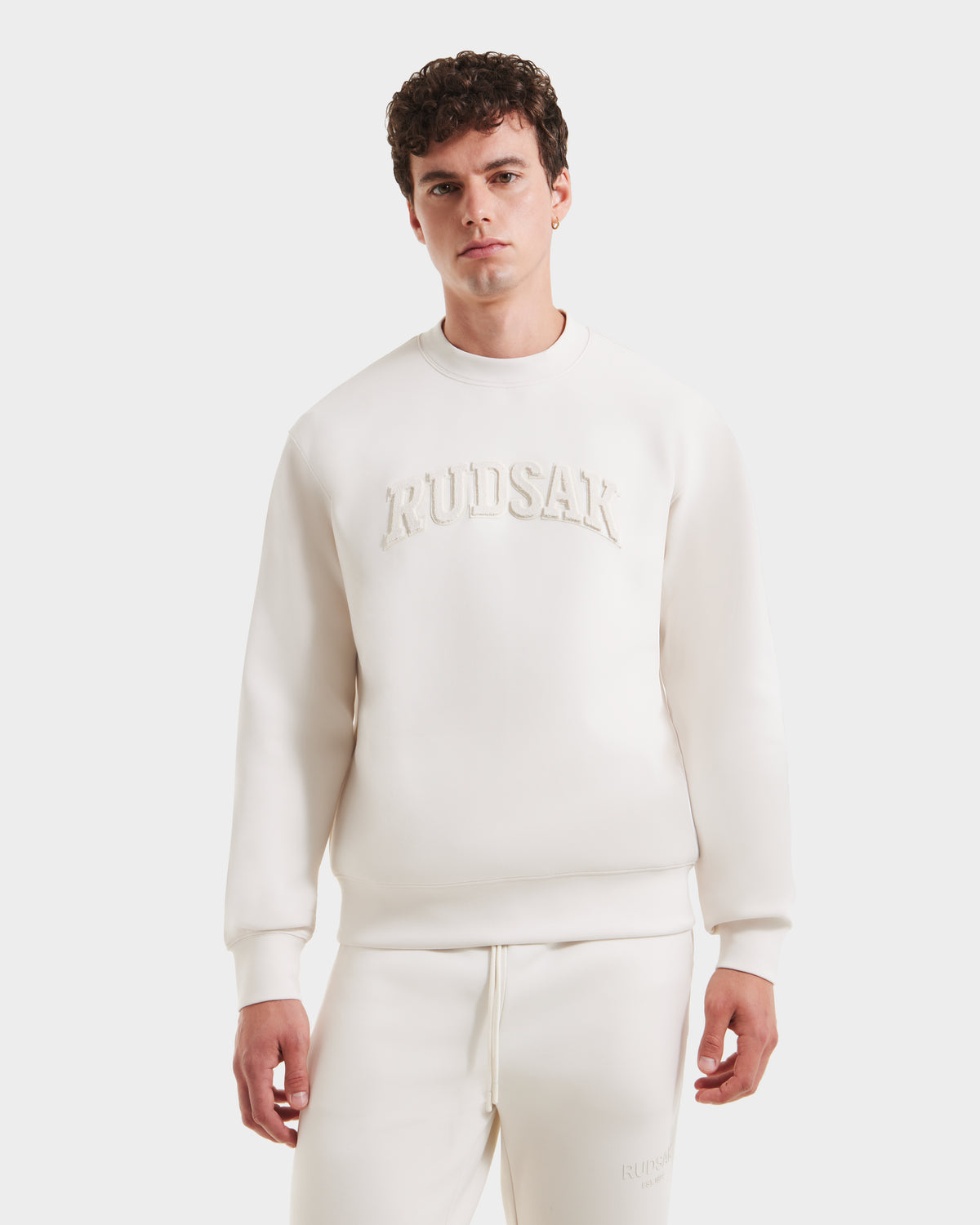 A young man with curly hair wears the Rudsak MILOS - 6525646 Vanilla men’s crewneck sweatshirt, crafted from a premium cotton blend and embossed with Rudsak on the chest, standing against a white background and looking at the camera.