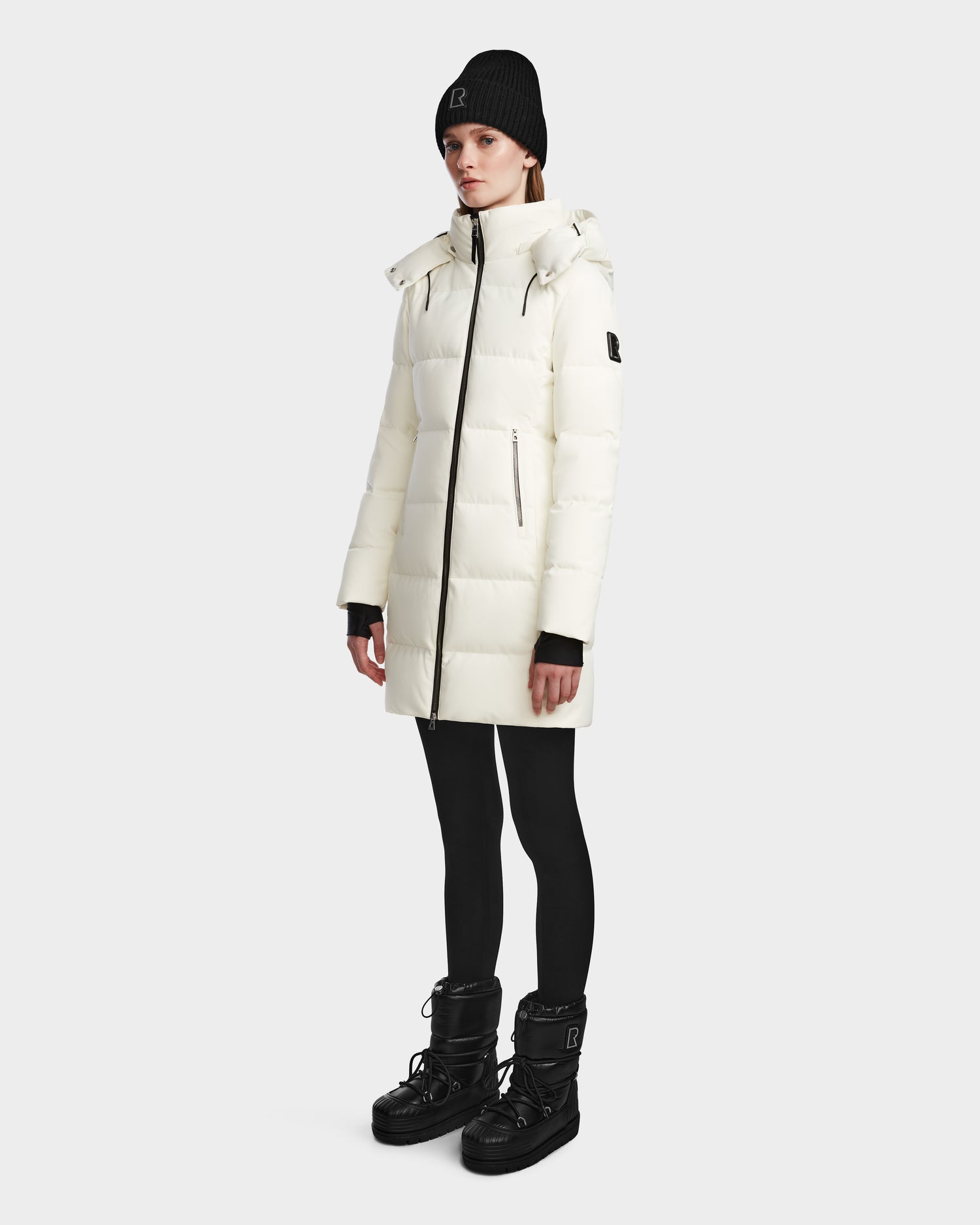 A person stands against a plain background, wearing the Rudsak MIRIAM - L123544 Iceland womens long white down puffer winter jacket with a high collar, zip pockets, sleeve logo, black leggings, black boots, and a black knit beanie.