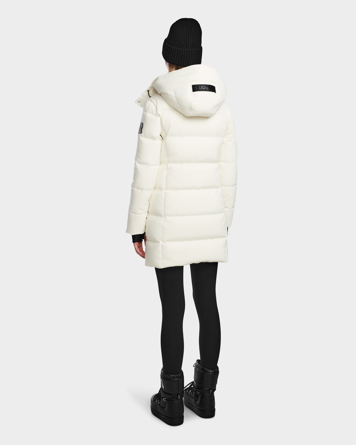 A person wearing the Rudsak MIRIAM - L123544 Iceland womens white down puffer coat with RDS-certified down, black leggings, boots, and a black knit beanie stands facing away from the camera against a plain light background.