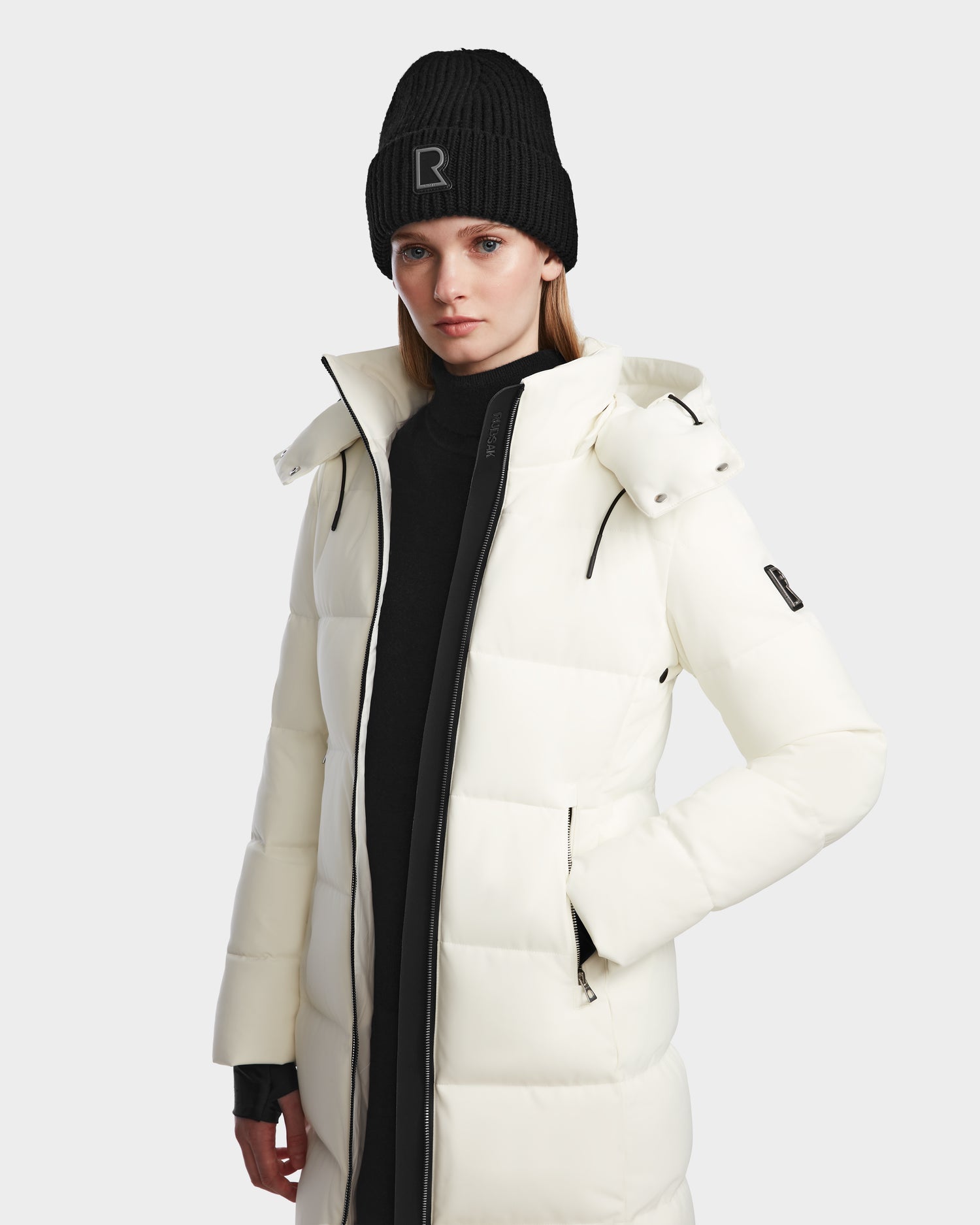 A woman wearing the Rudsak MIRIAM - L123544 Iceland white down puffer coat and a black beanie with an R stands against a plain light background, highlighting stylish winter essentials.