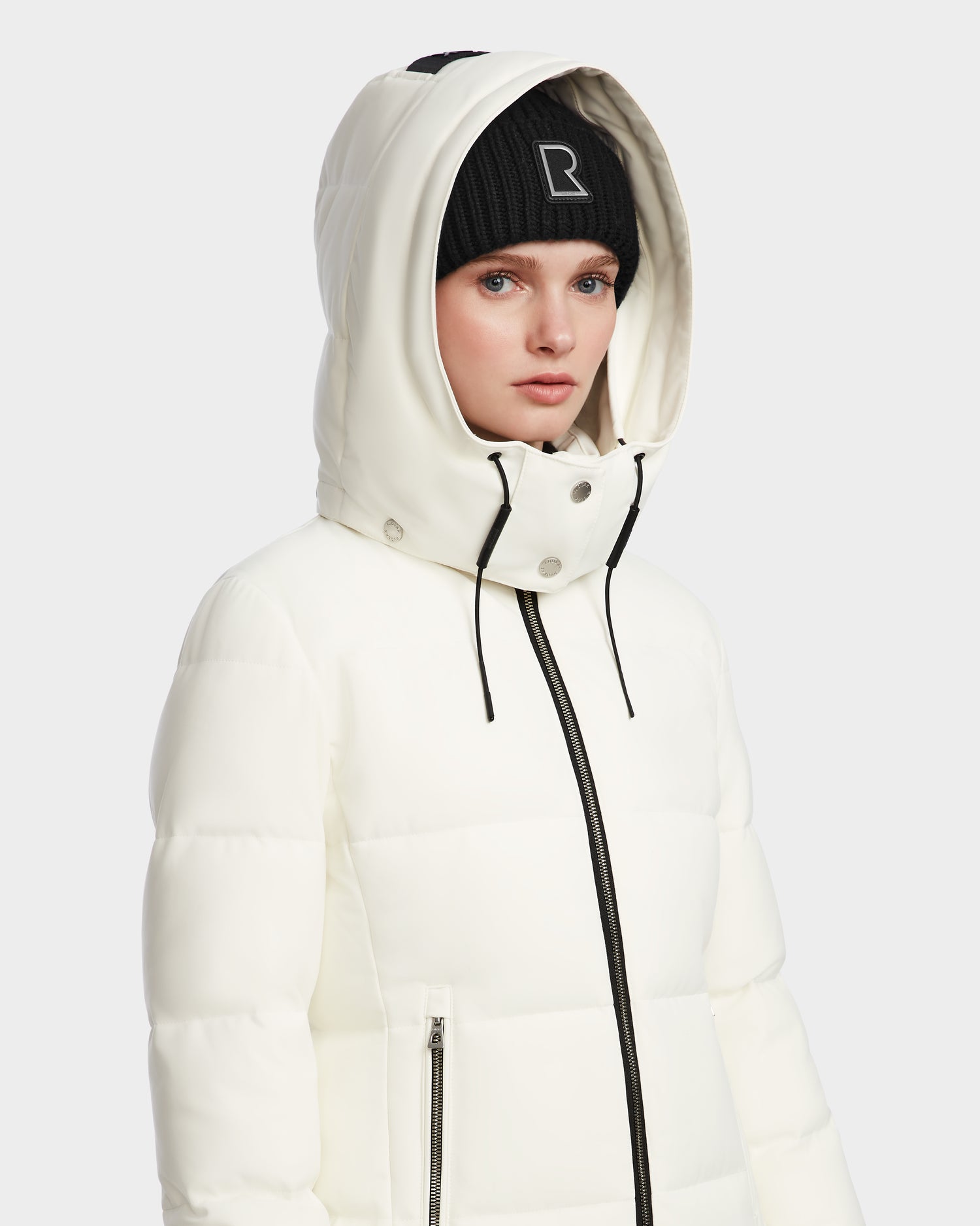 A woman wears the Rudsak MIRIAM - L123544 Iceland white RDS-certified down puffer jacket with the hood up and a black “R” logo beanie, facing forward with a neutral expression against a plain light background.