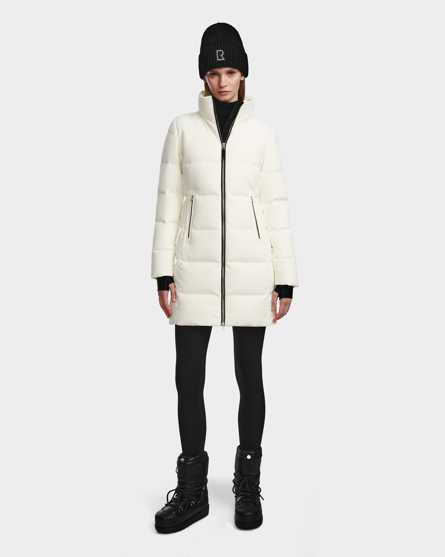 A person wears the Rudsak MIRIAM - L123544 Iceland white women’s down puffer, paired with a black beanie, black leggings, and black winter boots, standing facing forward against a plain light background.
