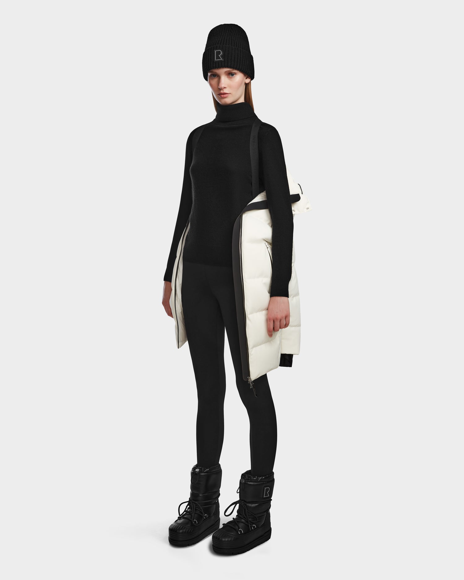 A woman stands against a plain background wearing a black turtleneck, leggings, beanie, and boots, with the Rudsak MIRIAM - L123544 Iceland women’s winter jacket draped over one shoulder.