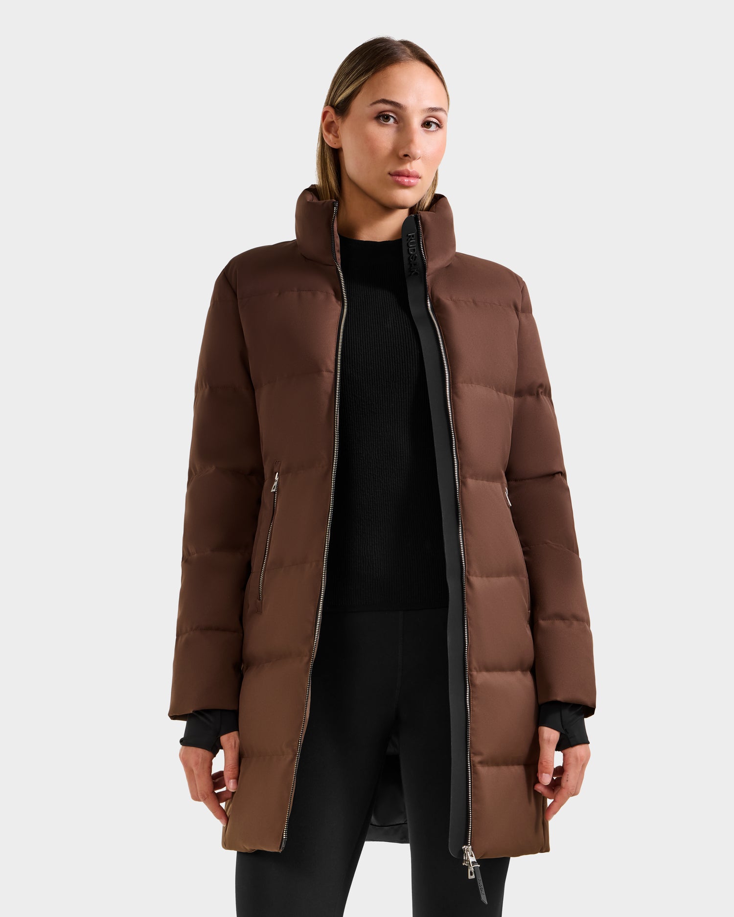 A woman faces forward wearing the Rudsak MIRIAM - L123544 Mocha women’s down puffer with a weather-resistant shell and zippered pockets over a black outfit, set against a plain light gray background—ideal for any winter wardrobe.