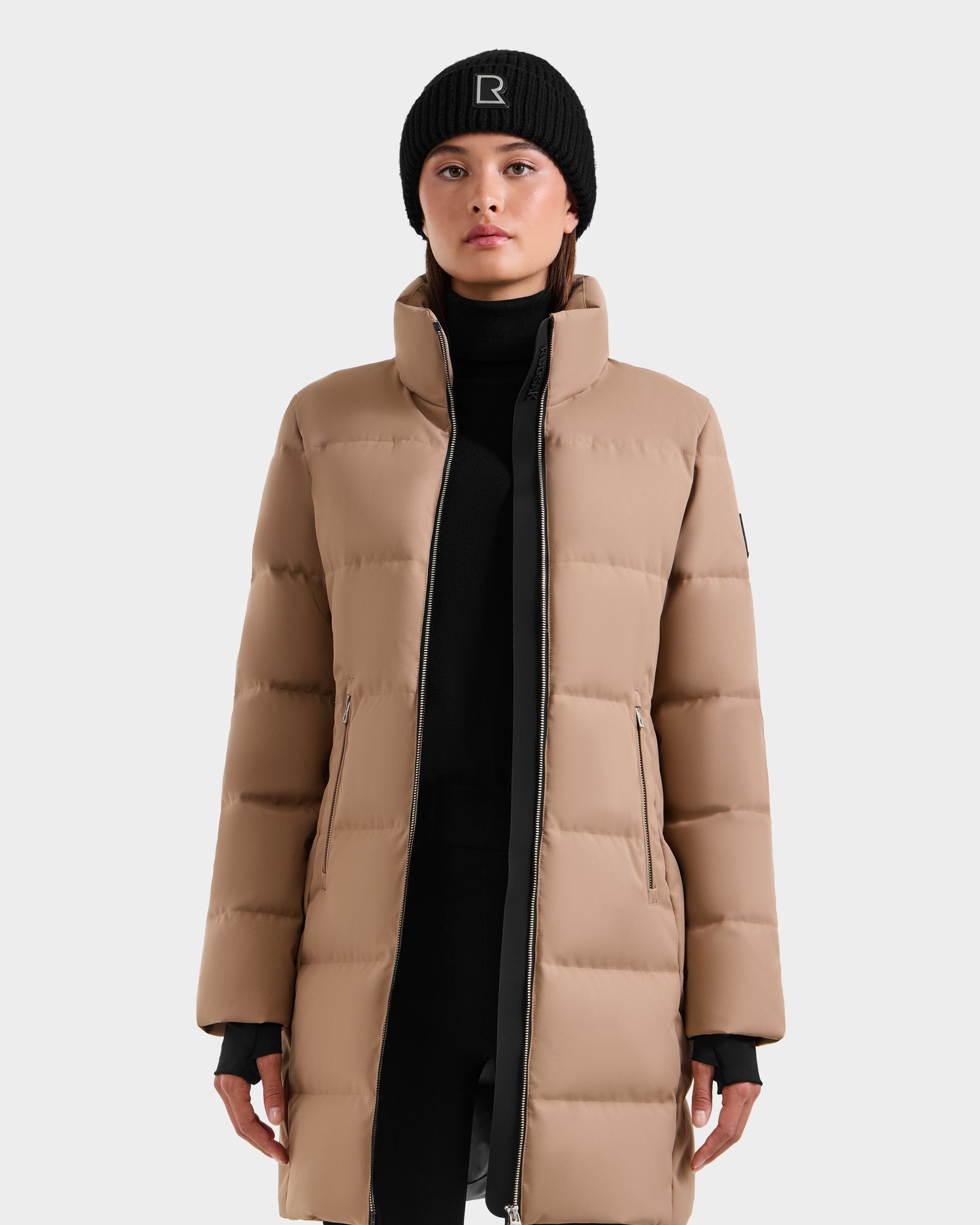 A person faces forward in the Rudsak MIRIAM - L123544 Sand women's down puffer coat with a high collar and front zipper, paired with a black beanie featuring a white R, against a plain light gray background.
