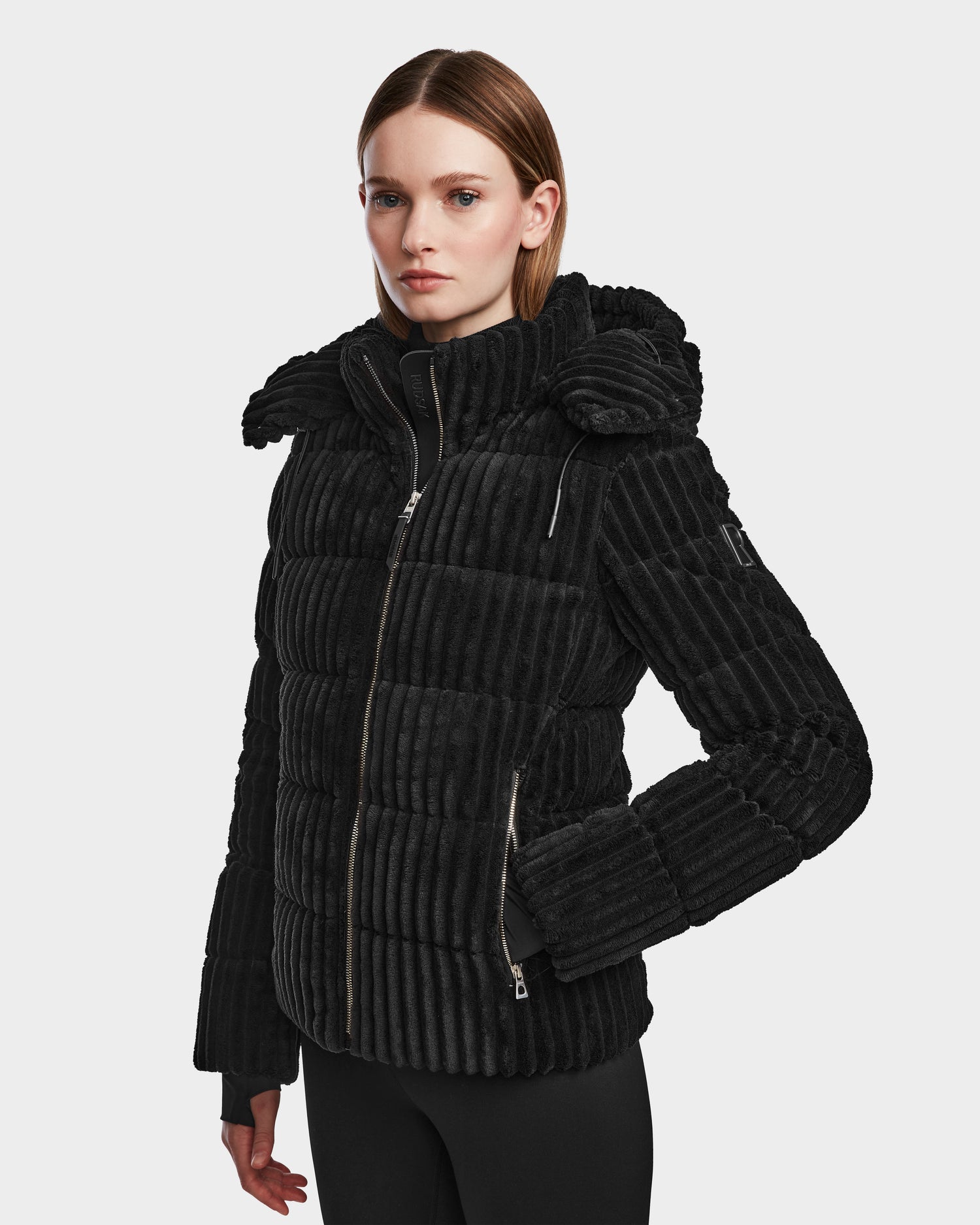 A woman with straight, shoulder-length hair wears the Rudsak MIRRY FF - L124507 Black women's short down puffer jacket, featuring a high collar and zippered pockets, against a plain light gray background.