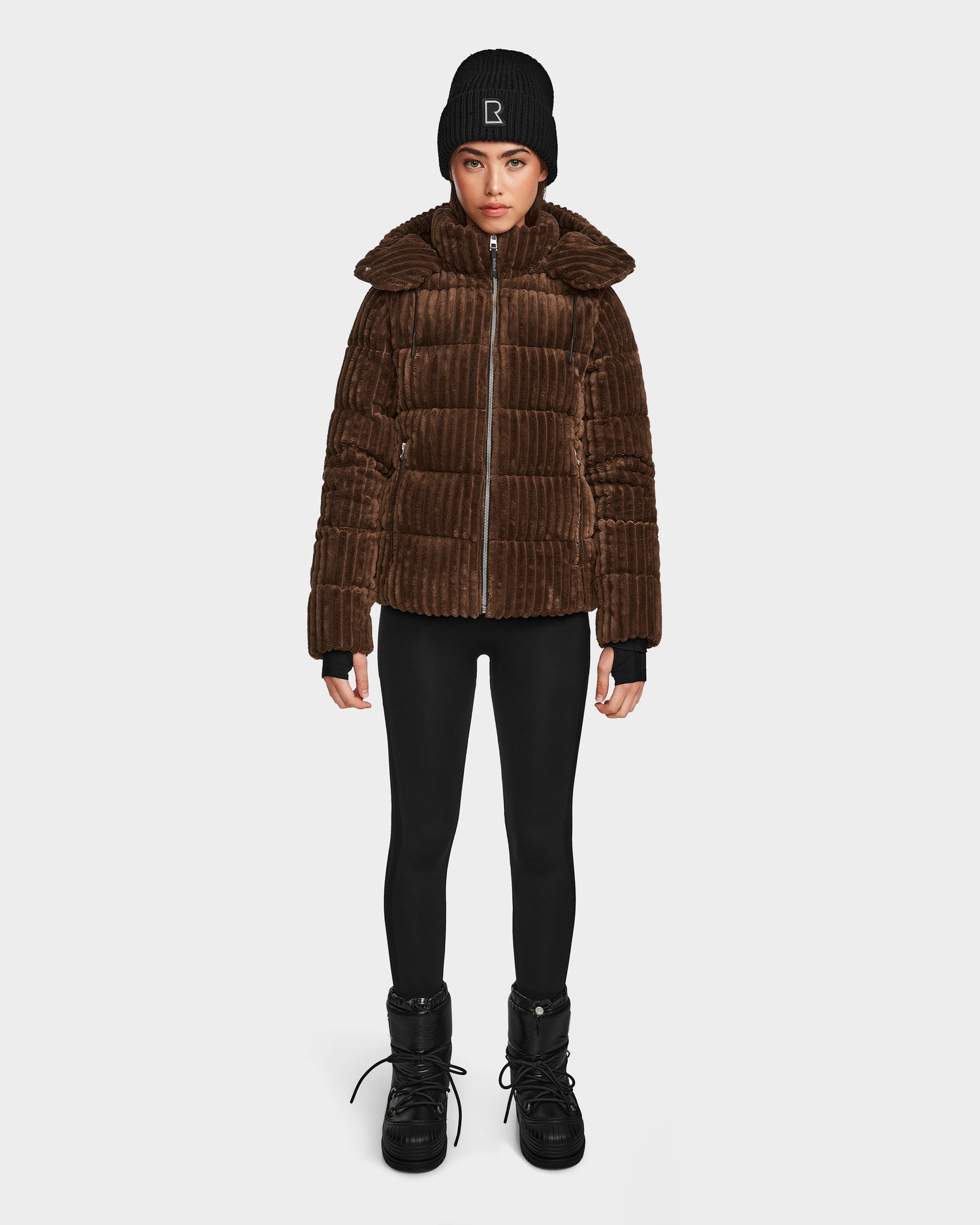 A person stands facing forward wearing the Rudsak MIRRY FF - L124507 Espresso women's short down puffer jacket, paired with black leggings, black lace-up boots, and a black beanie against a plain white background.