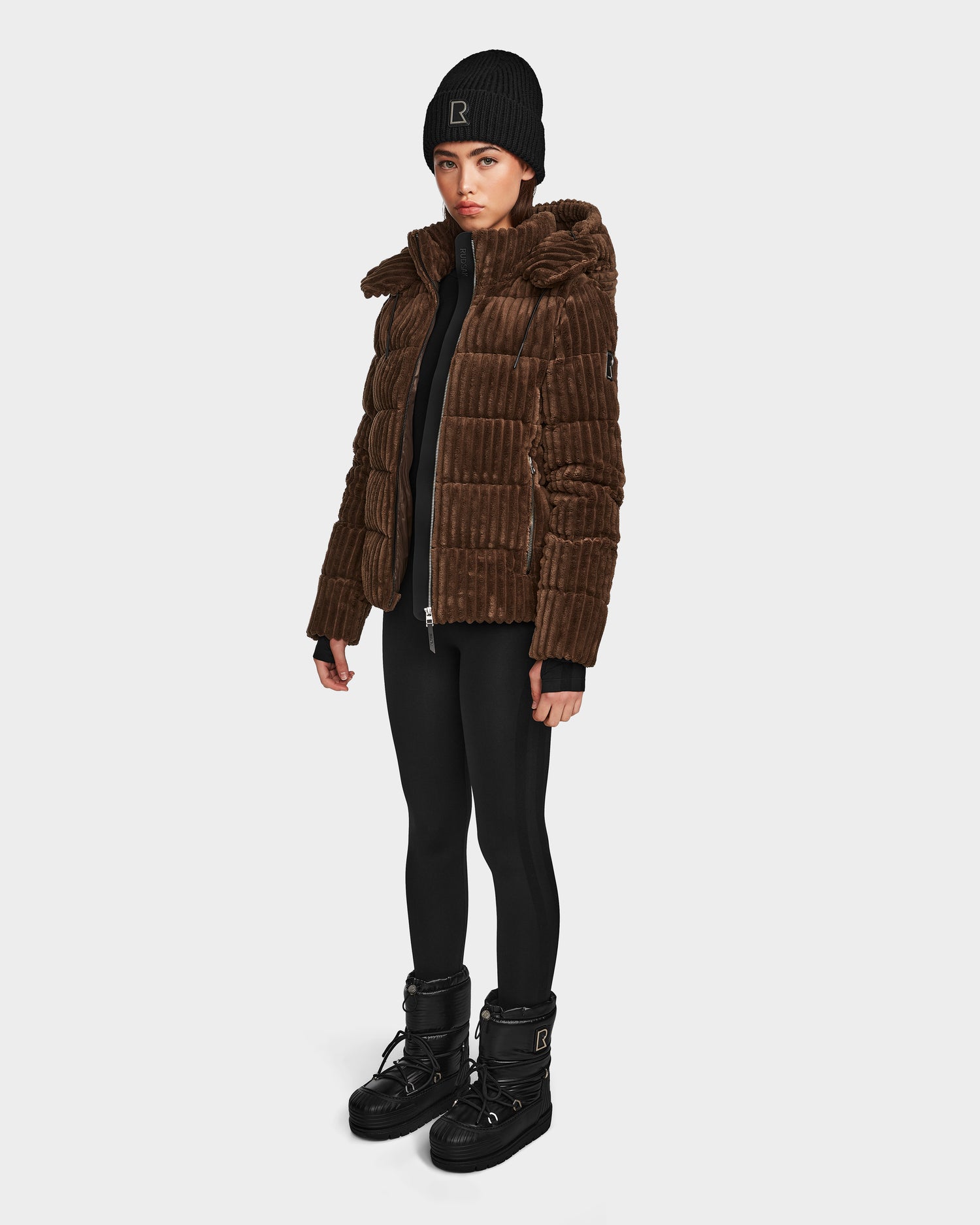 A woman stands against a white background wearing the Rudsak MIRRY FF - L124507 puffer jacket in Espresso, paired with black leggings, winter boots, and a black beanie. She looks forward with a neutral expression.