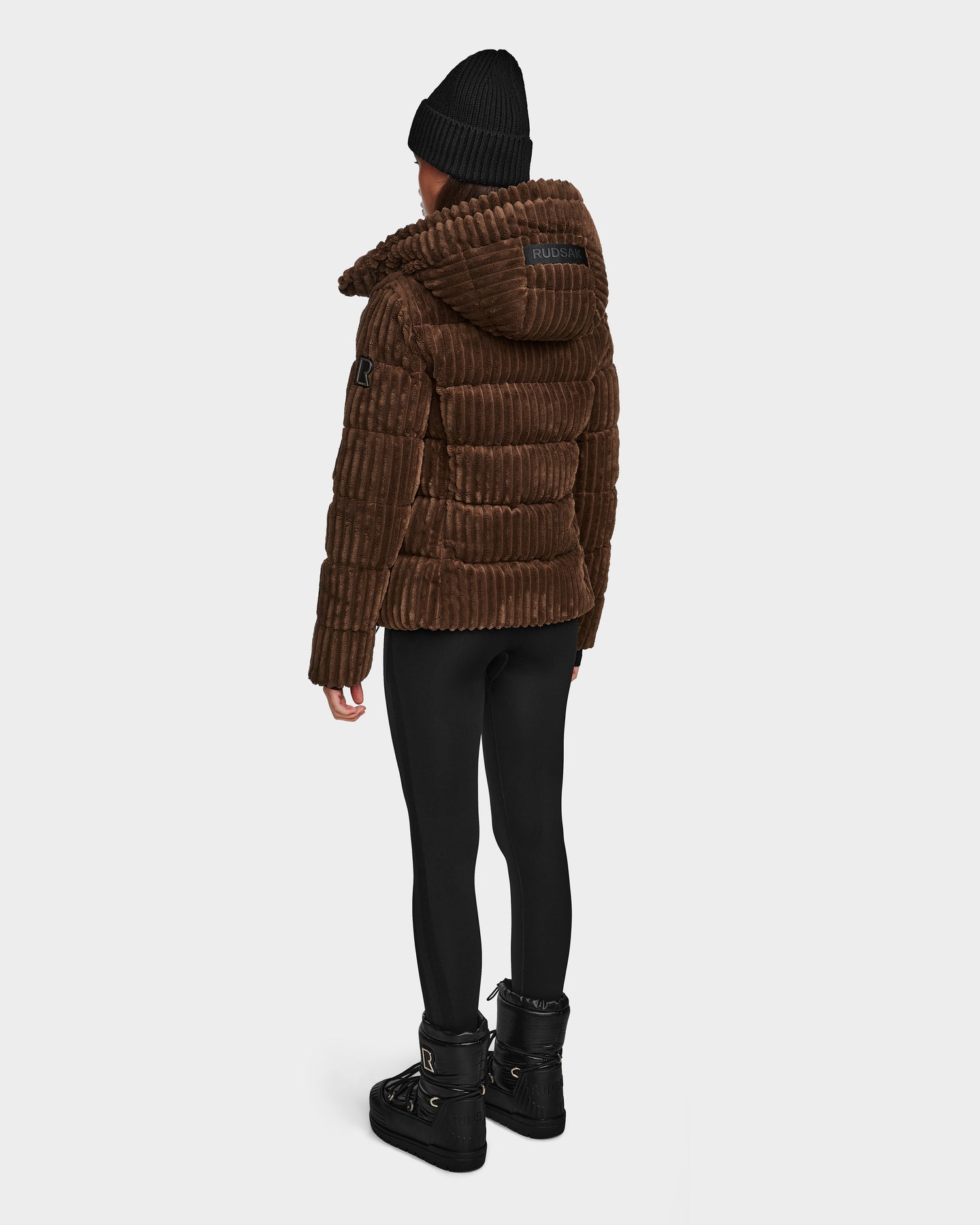 A person wears the Rudsak MIRRY FF - L124507 Espresso velvet corduroy puffer jacket, paired with a black beanie, leggings, and boots, standing with their back to a plain white background.